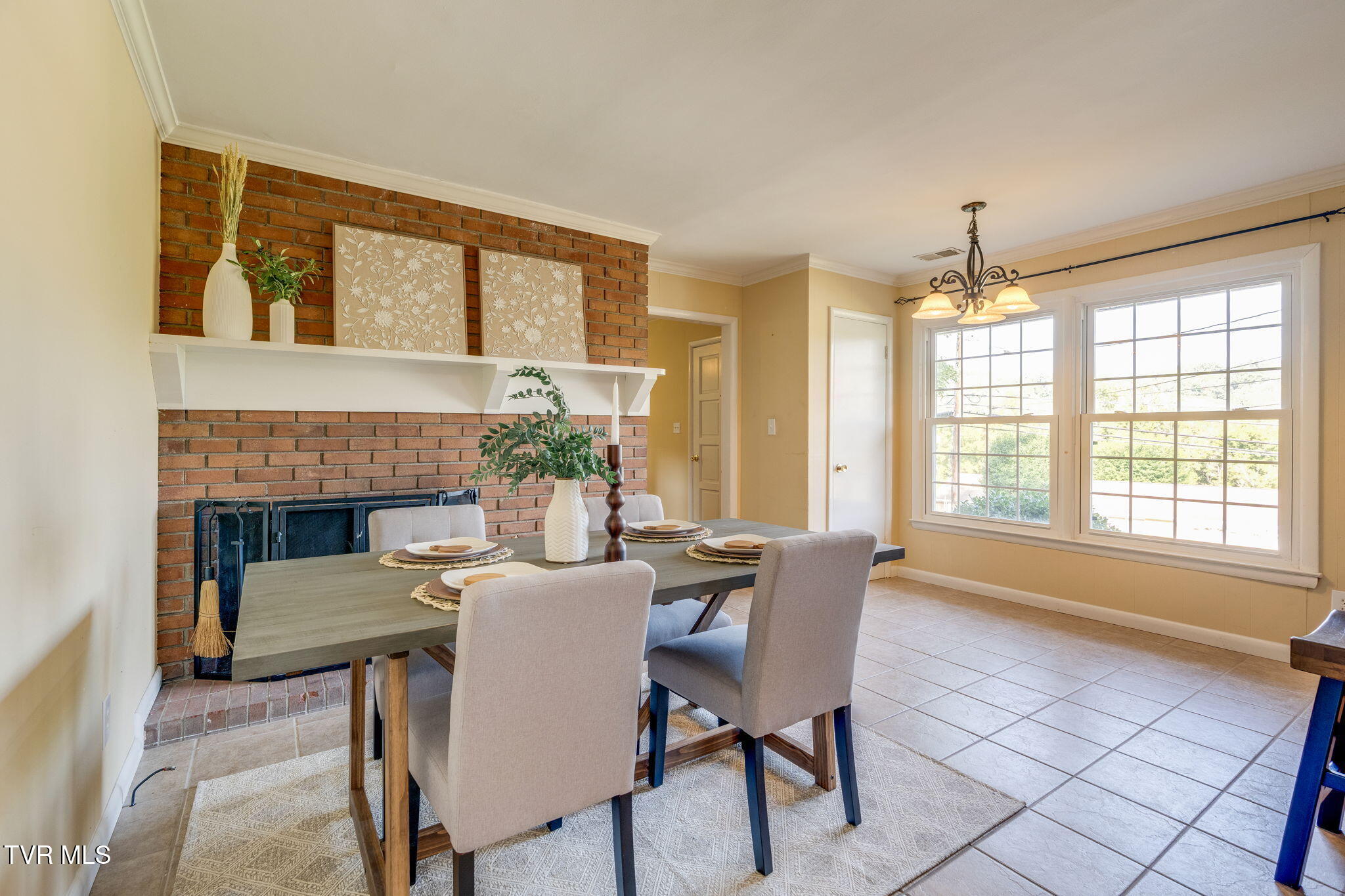 Dining room with a brick fireplace, staged table, upholstered chairs, and large sunlit windows.