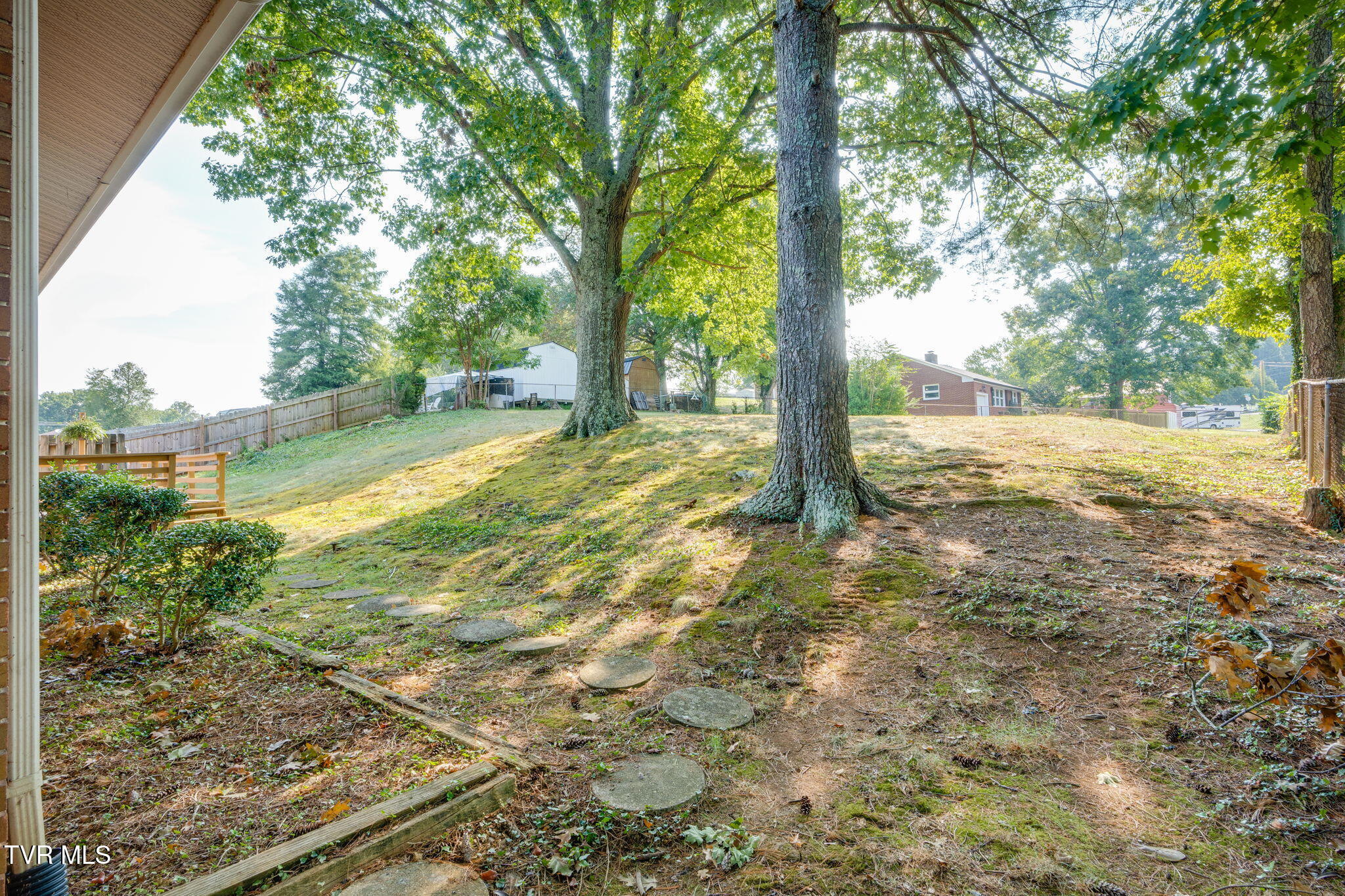 Sloping backyard with mature shade trees, stepping stones, shrubs and a wooden fence.
