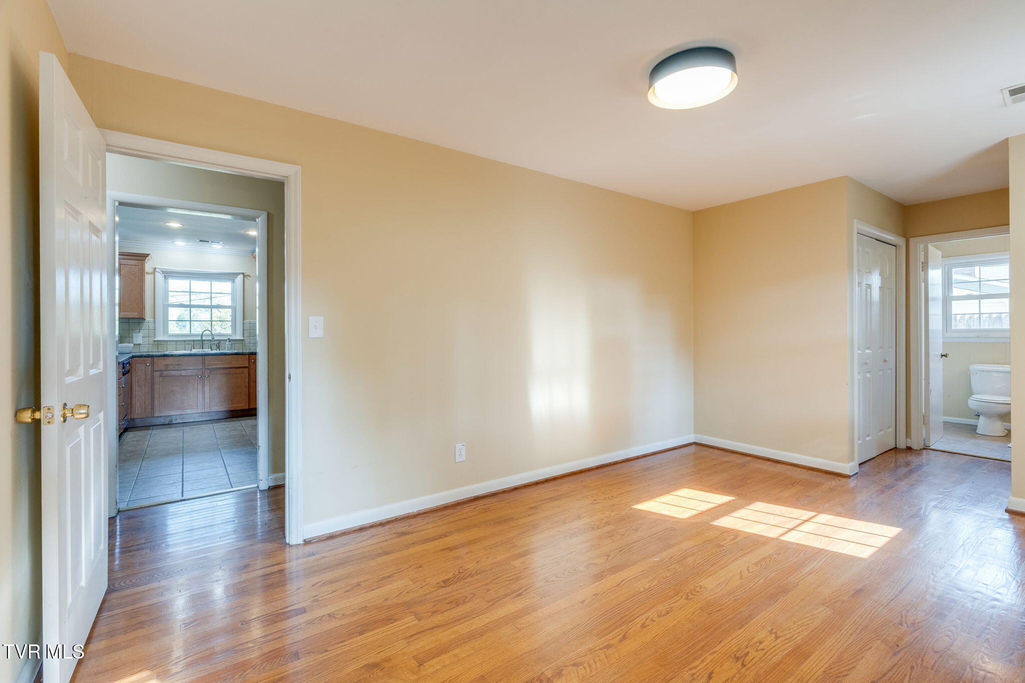 Empty room with polished hardwood floors, a kitchen doorway, and a bathroom doorway.