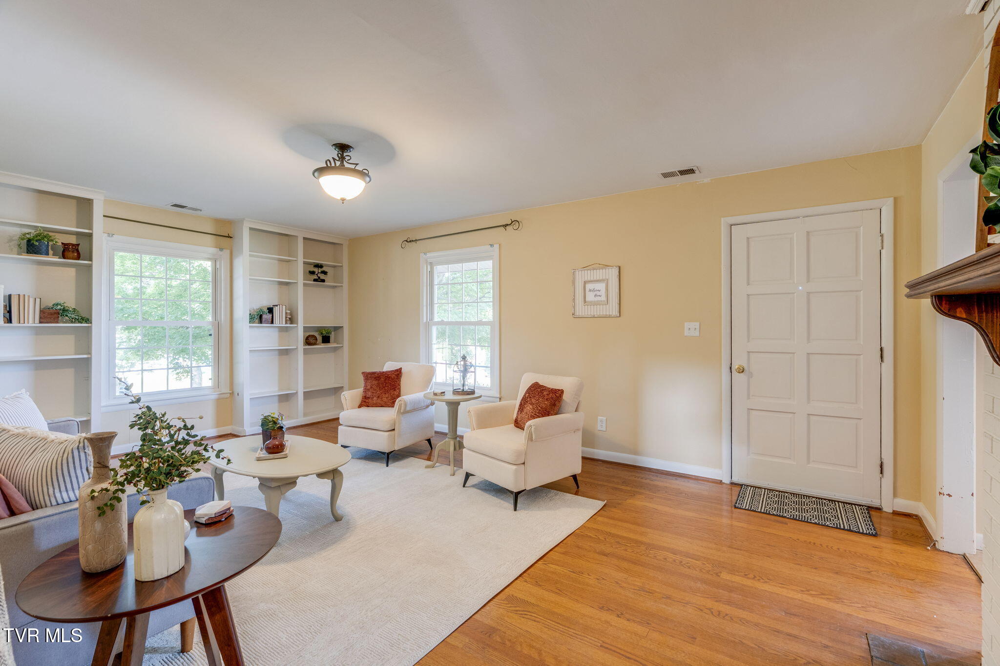 Staged living room with built-in shelving, cream armchairs, a large rug, and hardwood floors.