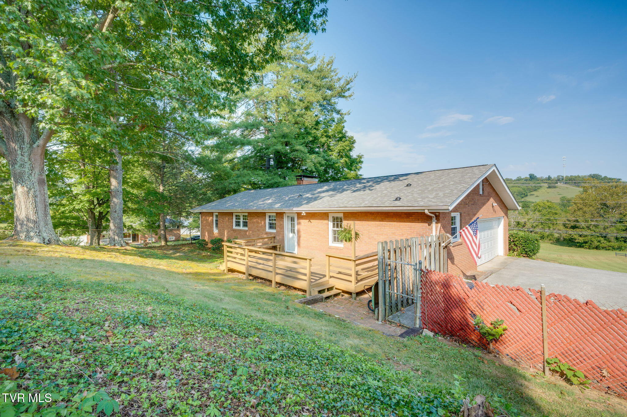Brick ranch home with a wooden deck, attached garage, and mature trees on a sloping lot.