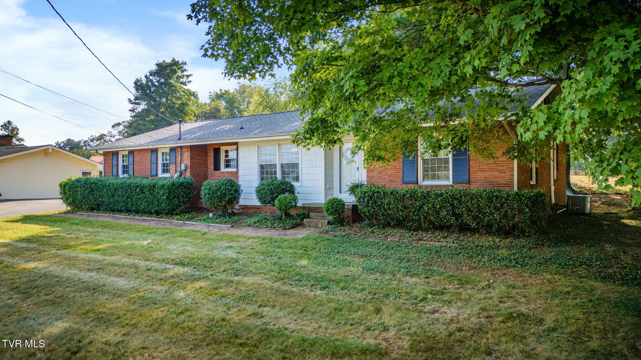 Single-story brick house with white siding, blue shutters, and a large shade tree over the front lawn.