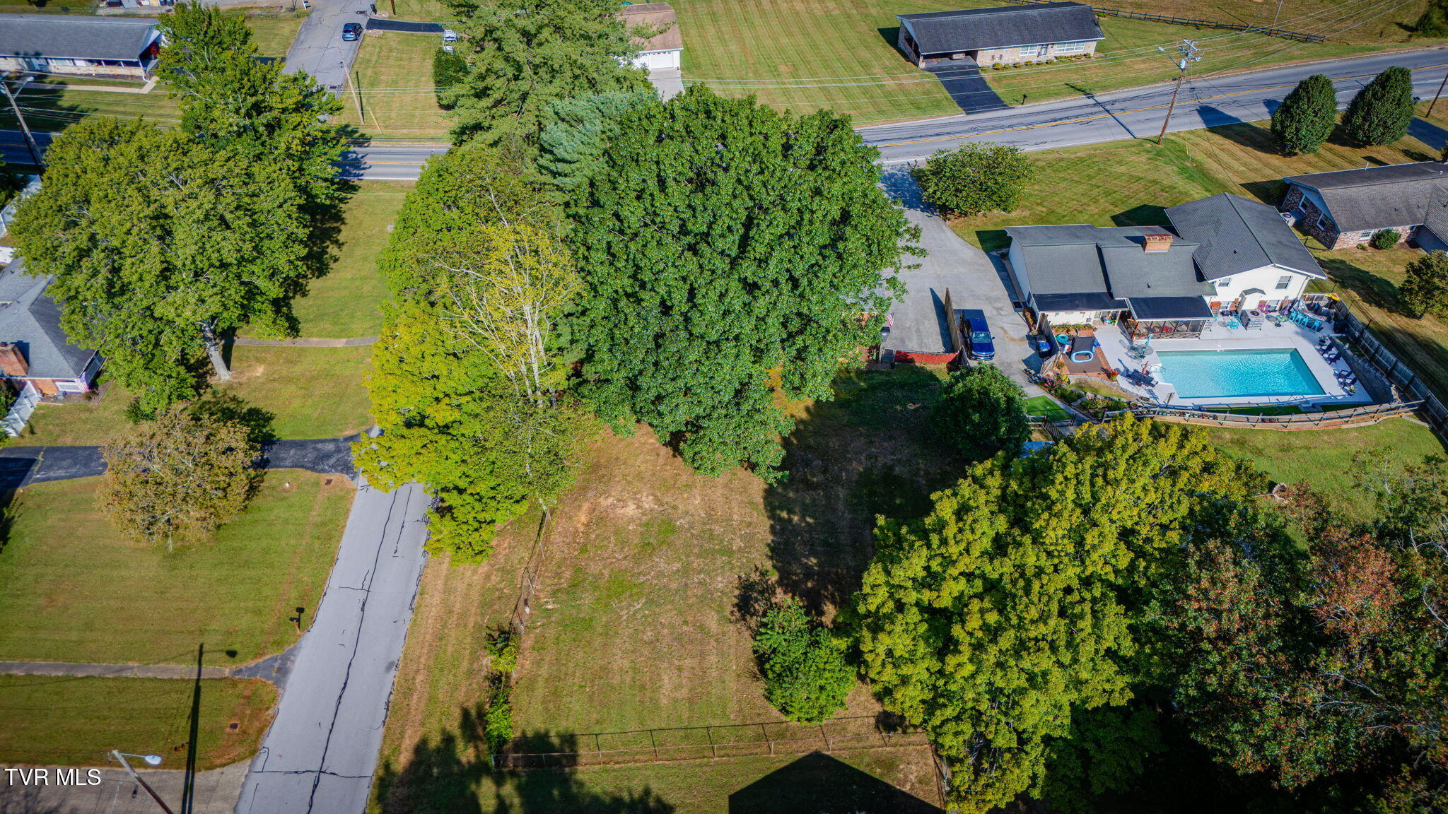 Aerial view of a home with a fenced pool, spacious tree-lined yard, and neighboring houses.