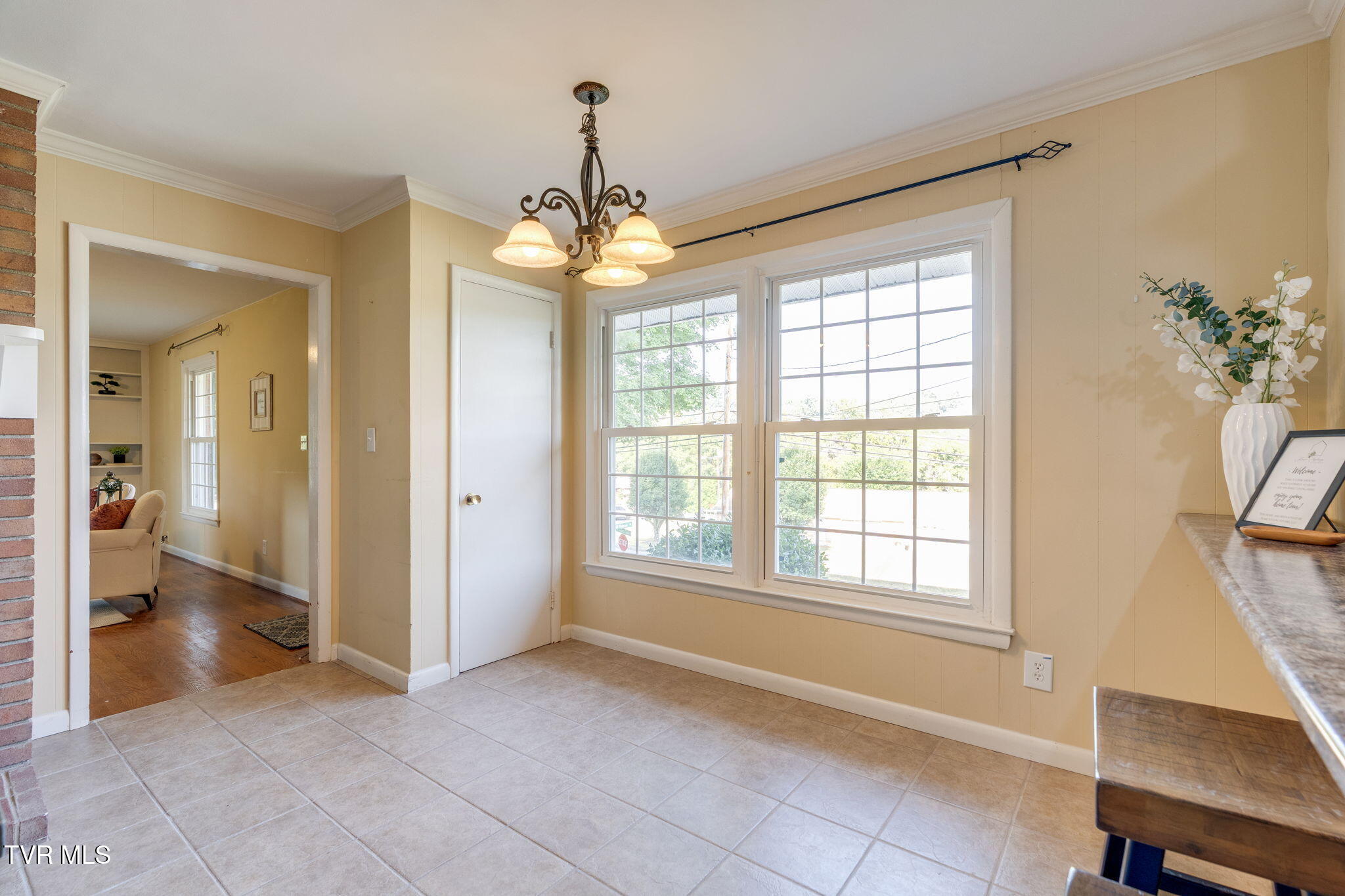 Dining area with tiled floor, chandelier, and two large windows overlooking greenery.