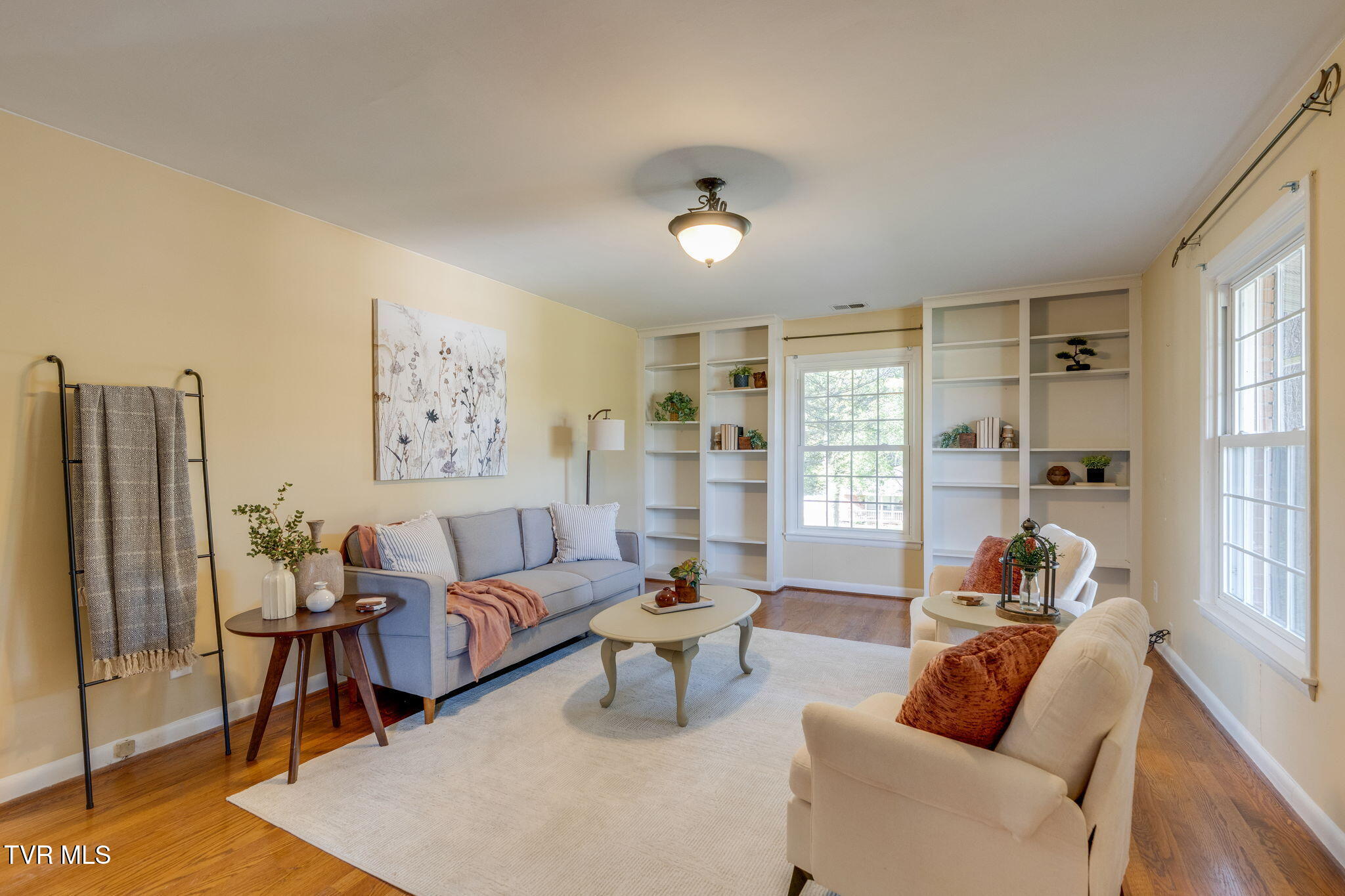 Staged living room with built-in bookshelves, a gray sofa, cream armchairs, and large windows.