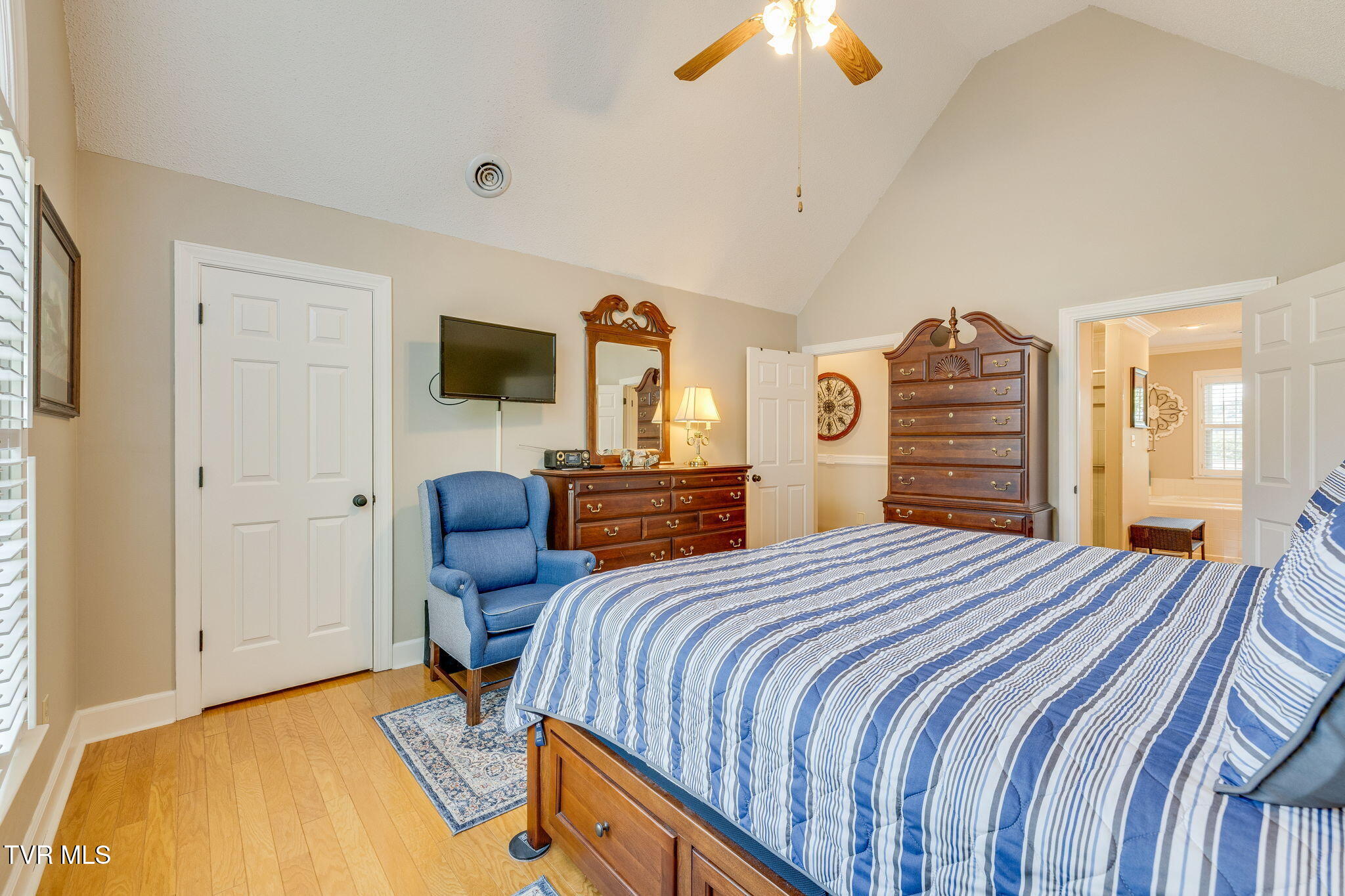 Furnished bedroom with a striped blue bed, blue armchair, and wooden dressers beside an adjoining bathroom.