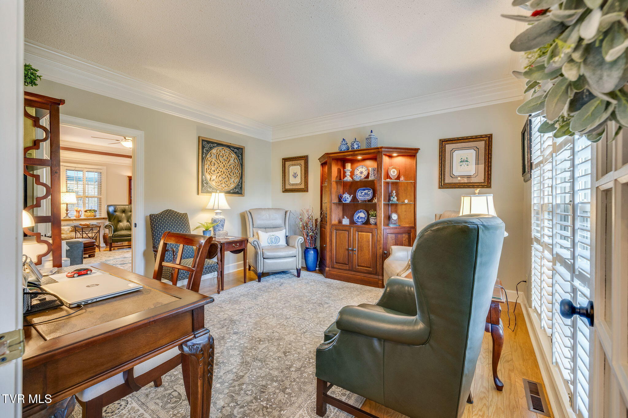 Furnished study with a wooden desk, display cabinet, upholstered chairs, and plantation shutters.