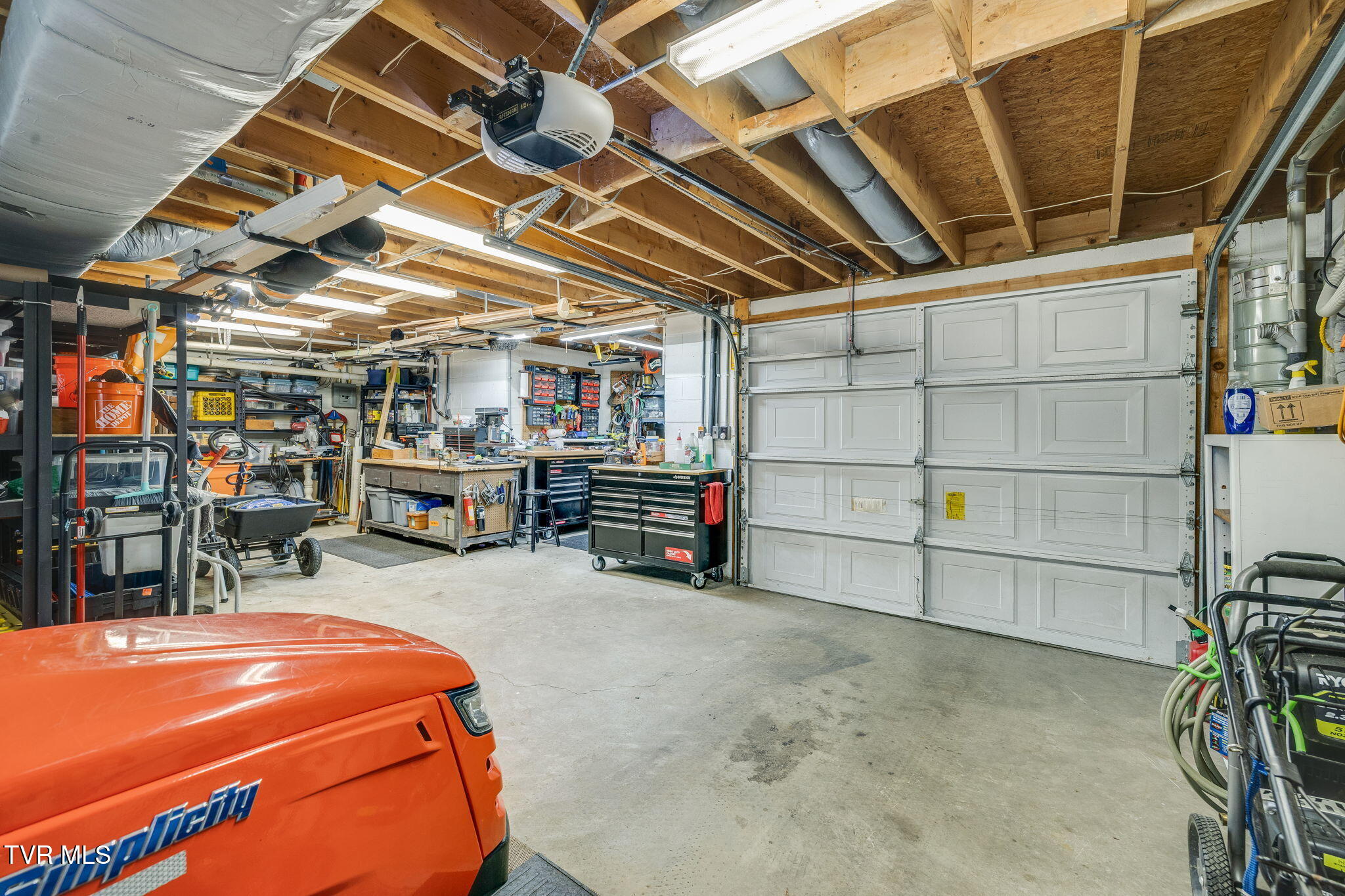 Garage workshop with exposed ceiling joists, extensive tool storage, workbenches, and an orange riding mower.