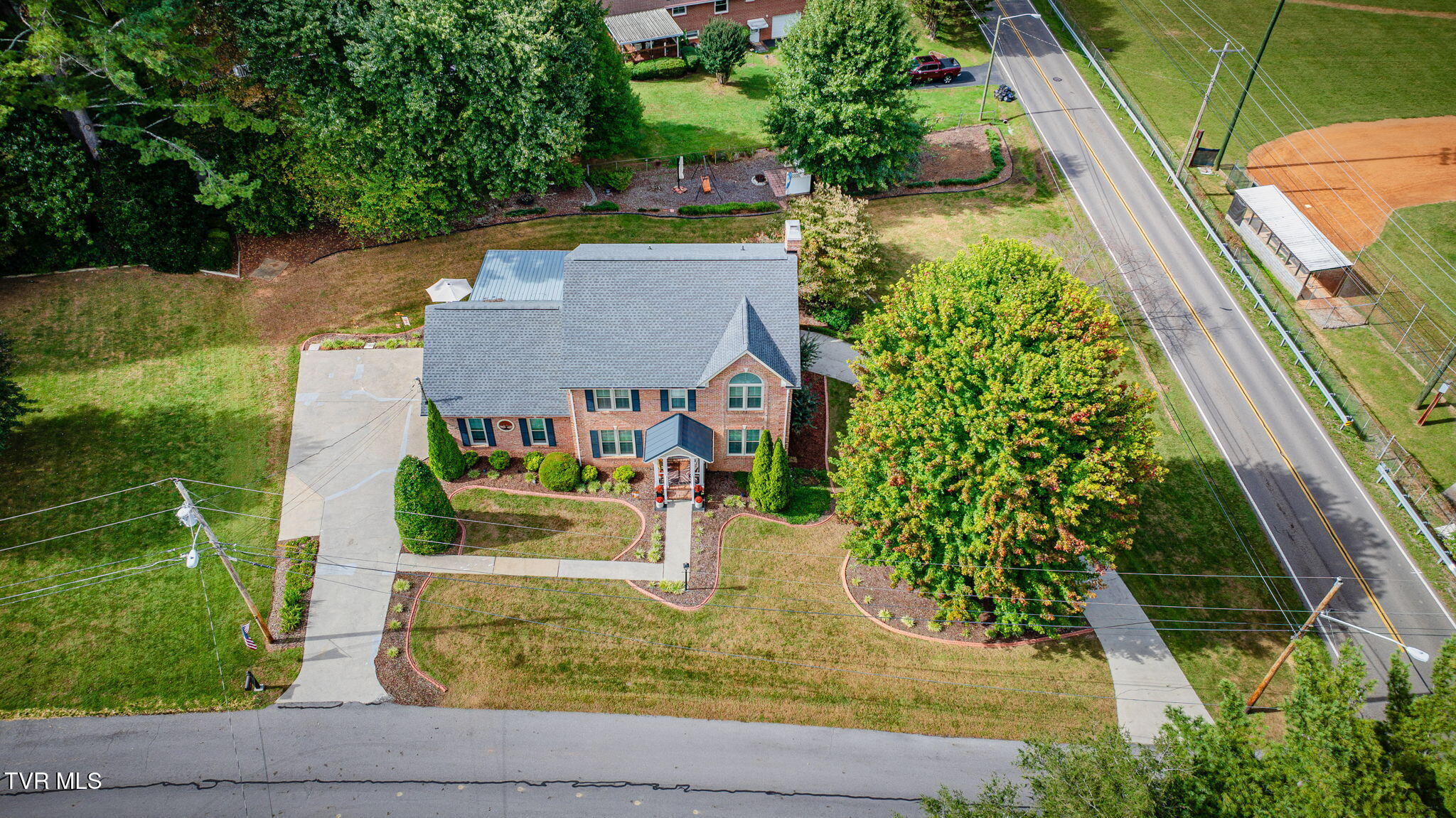Two-story brick house with landscaped yard and driveway, seen from above beside a road.