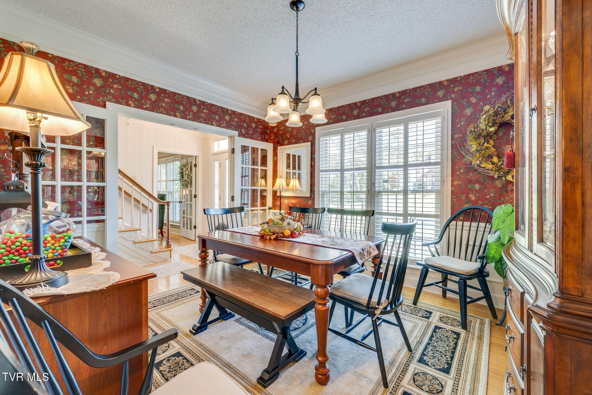 Dining room with floral red wallpaper, a dark wood table, black chairs, and bright shuttered windows.
