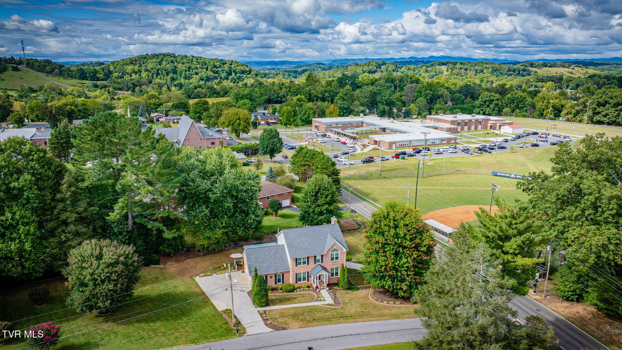 Brick two-story home with a landscaped yard, viewed aerially with a school and wooded hills beyond.