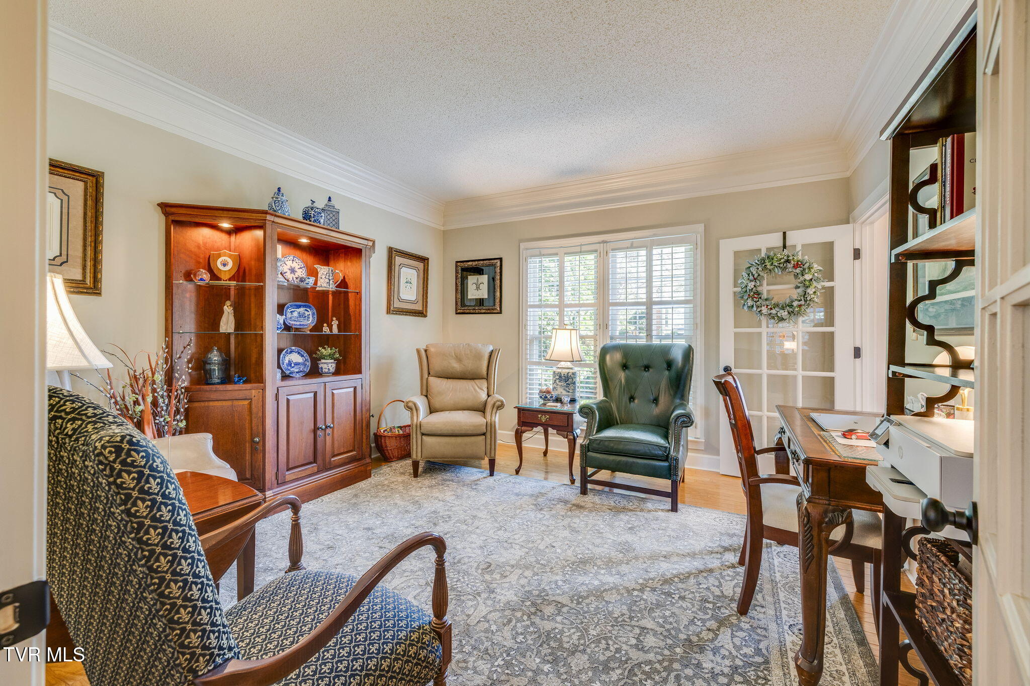 Furnished home office with a wooden display cabinet, two armchairs, and shuttered windows.