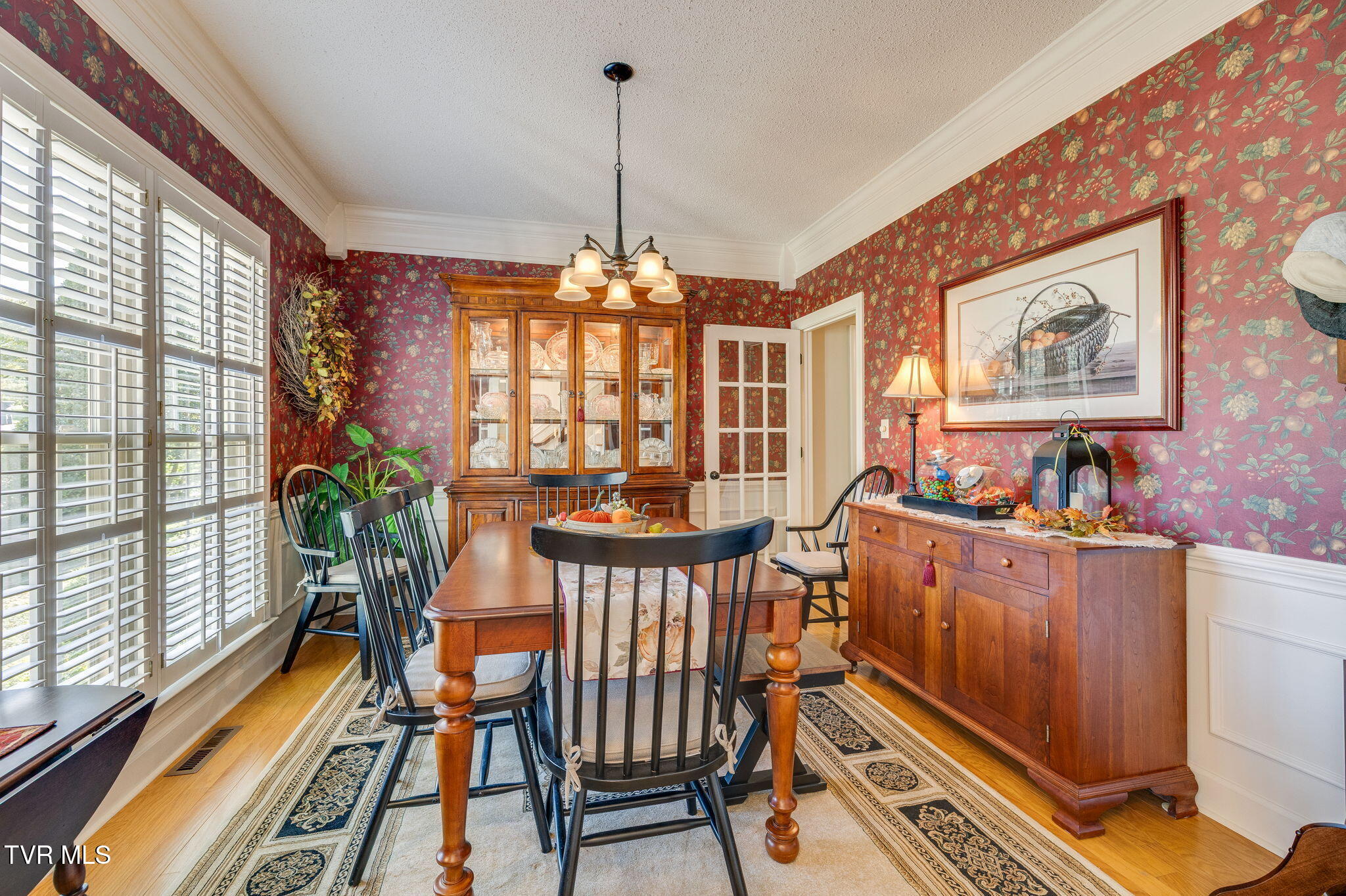 Dining room with red floral wallpaper, plantation shutters, and a wooden china cabinet.