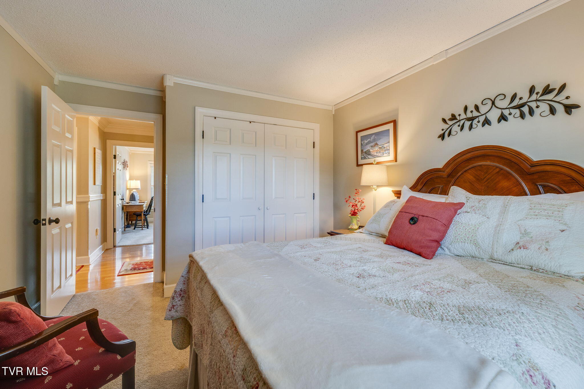 Bedroom with a carved wooden headboard, white floral bedding, red accent pillow, and open doorway.