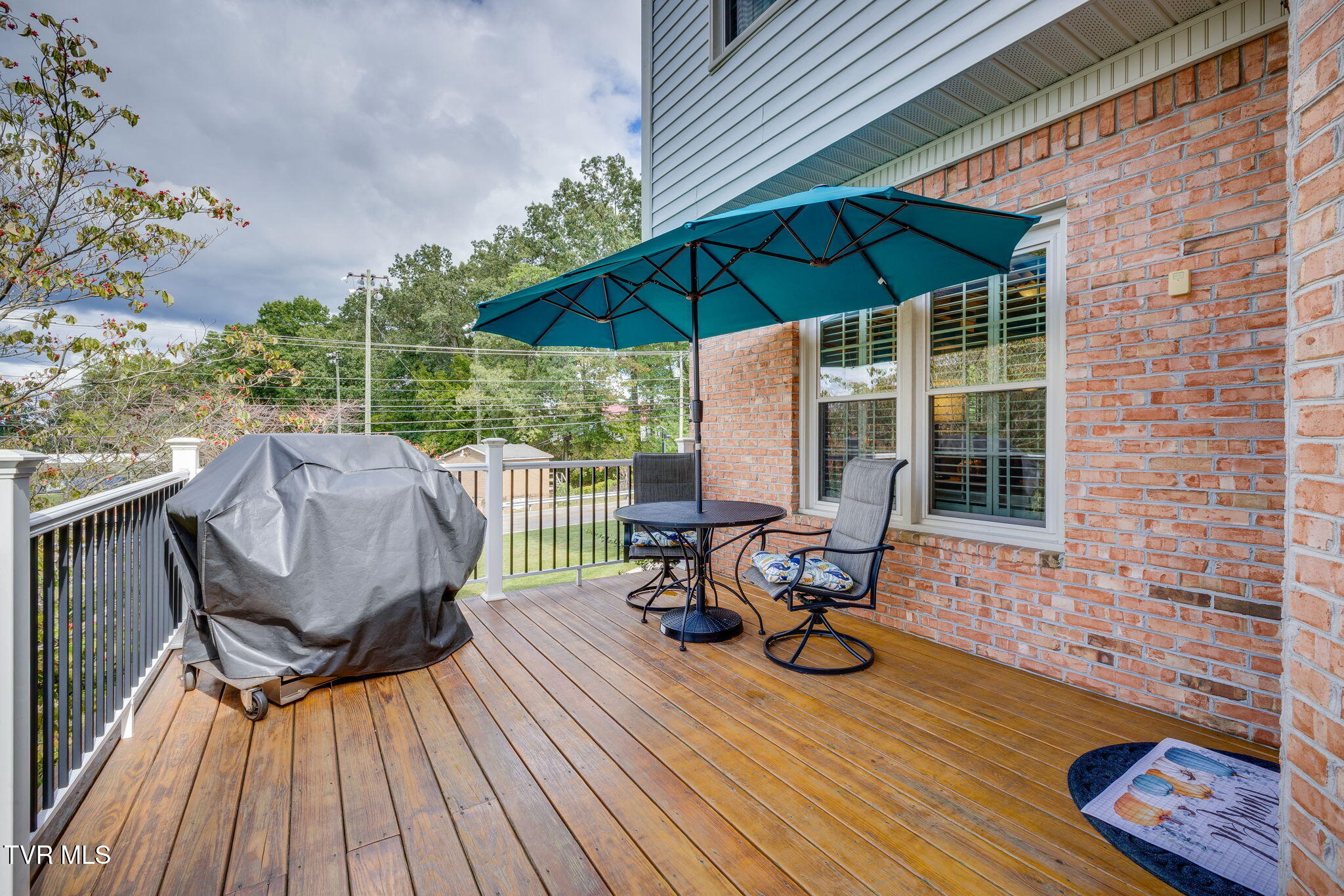 Wooden deck with a teal umbrella, round patio table, chairs, and covered grill beside a brick house.