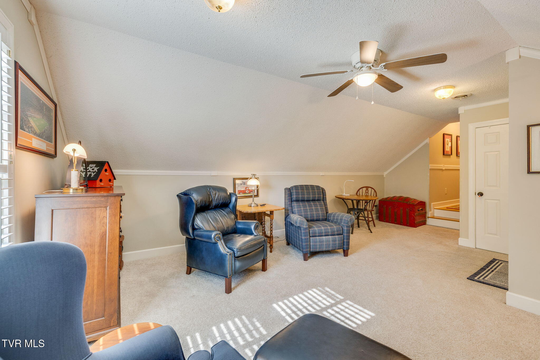 Upstairs sitting room with a sloped ceiling, blue armchairs, and sunlight across the carpet.