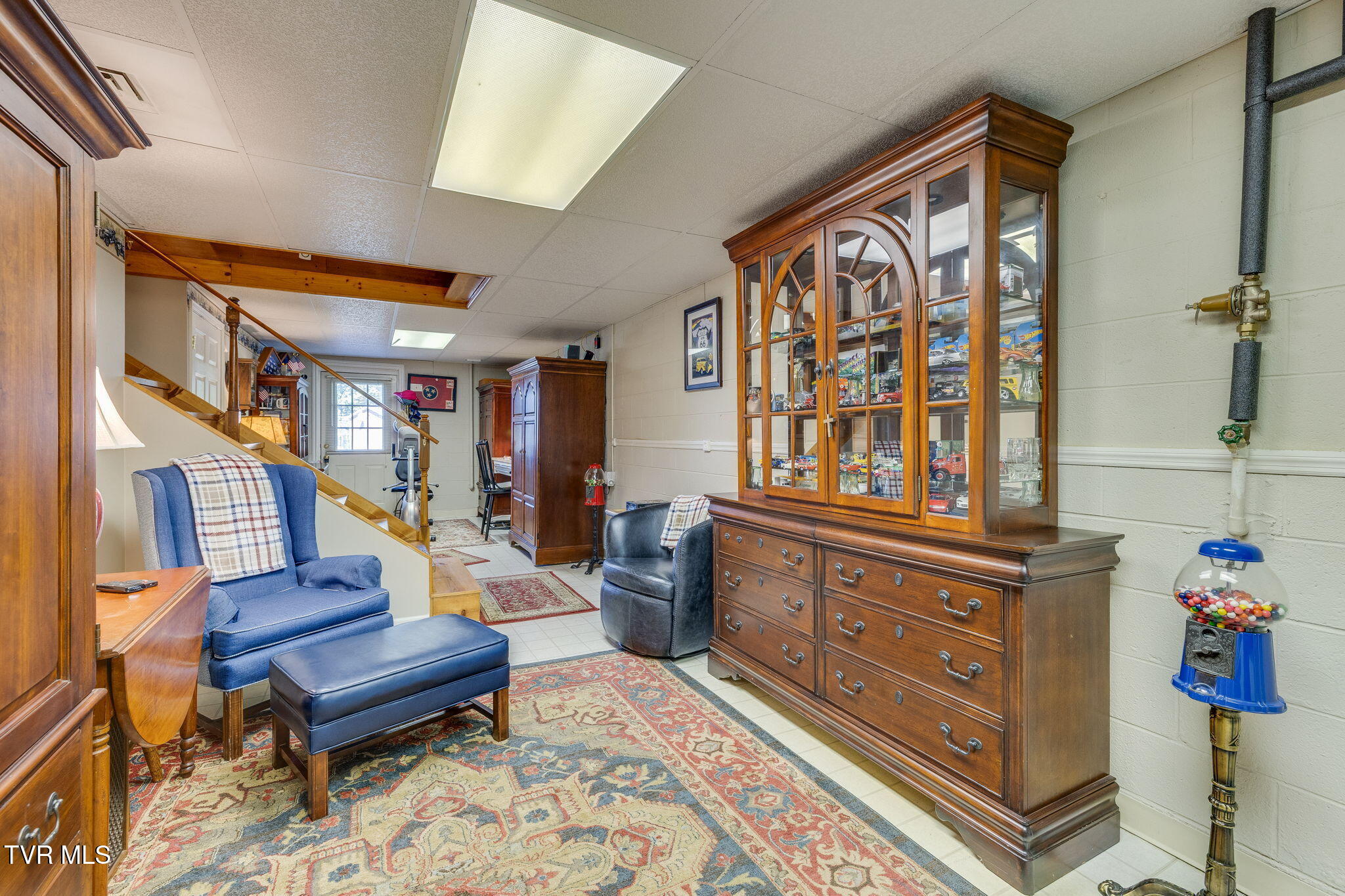 Furnished basement den with a large wooden display cabinet, model vehicles, staircase, and patterned rugs.