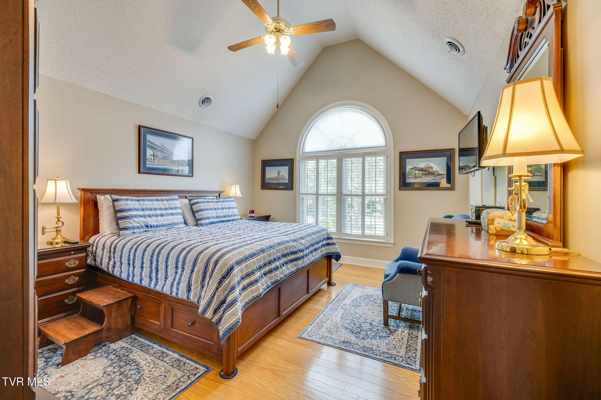 Furnished bedroom with a vaulted ceiling, arched shuttered window, and large wooden bed with blue striped bedding.