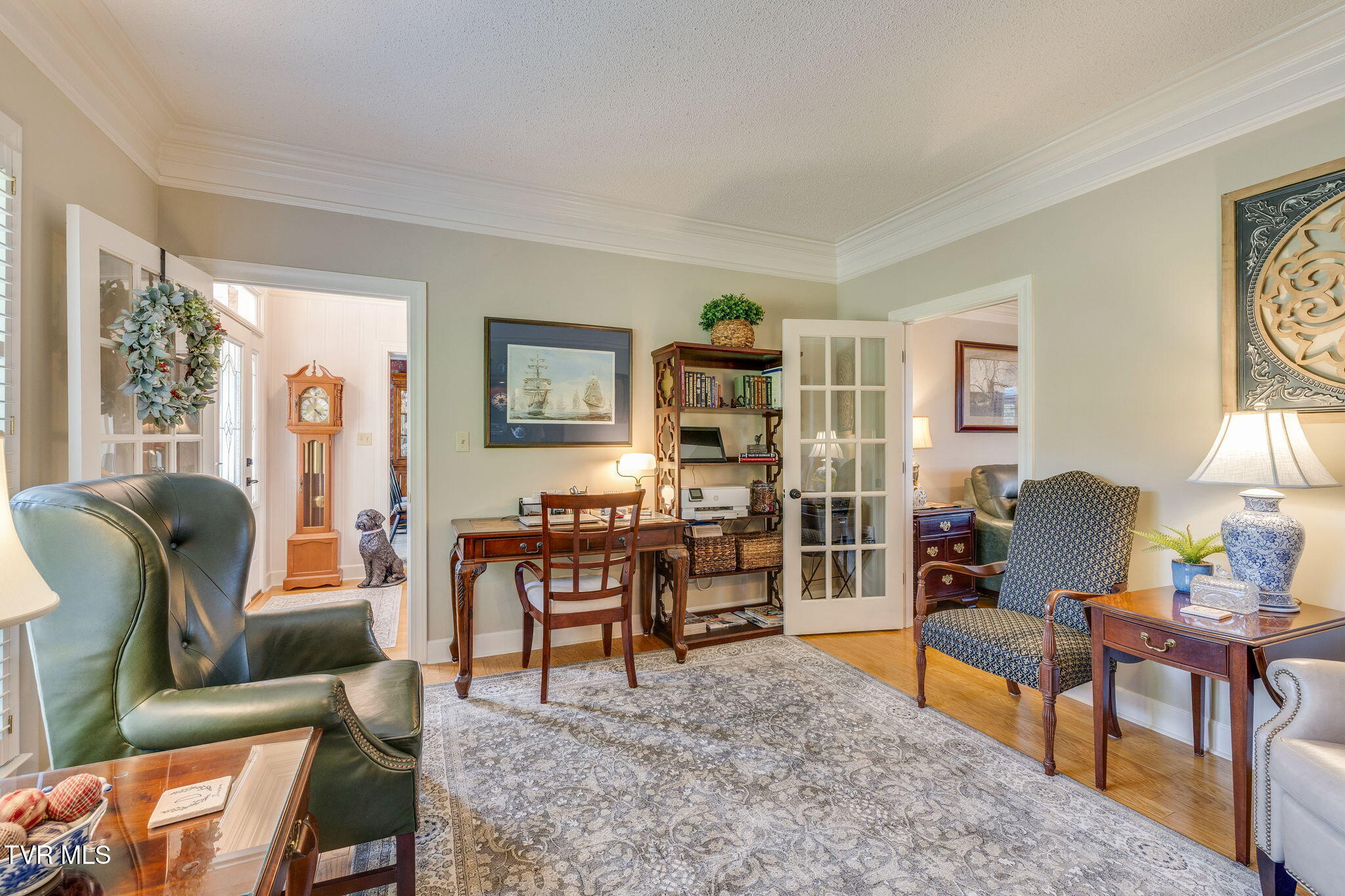 Furnished home office with a wooden desk, bookcase, green leather armchair, and French doors.
