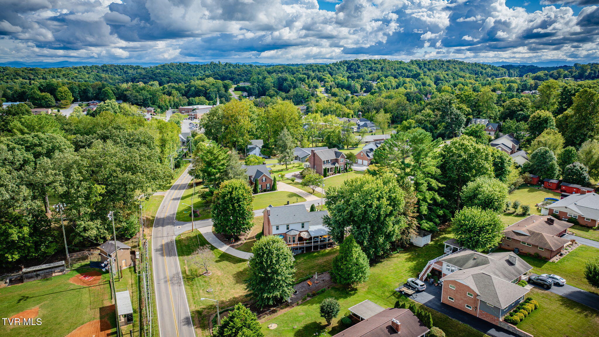 Aerial view of a wooded residential neighborhood with homes, a winding road, and distant mountains.