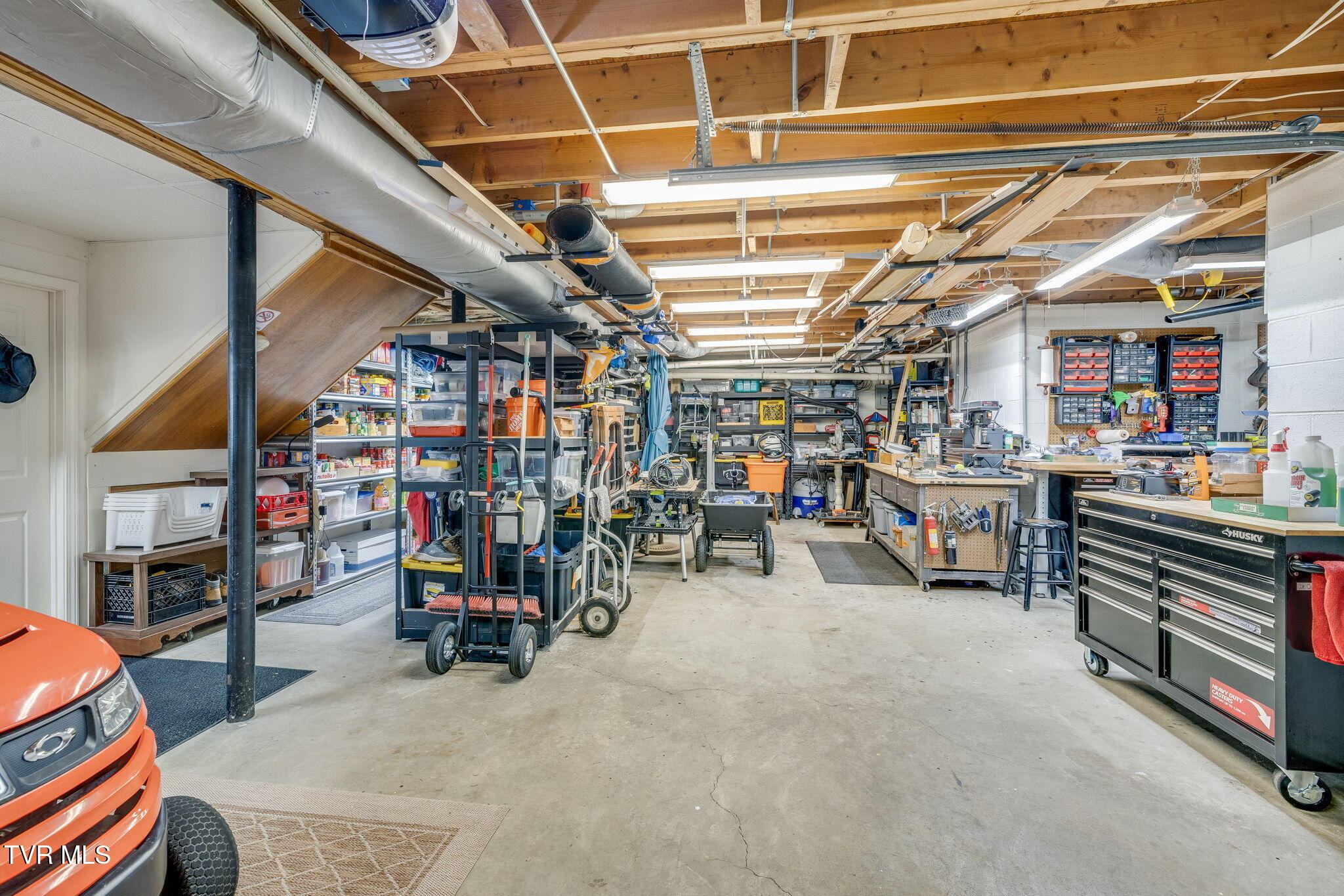 Spacious garage workshop with exposed beams, organized tool storage, workbenches, and an orange riding mower.