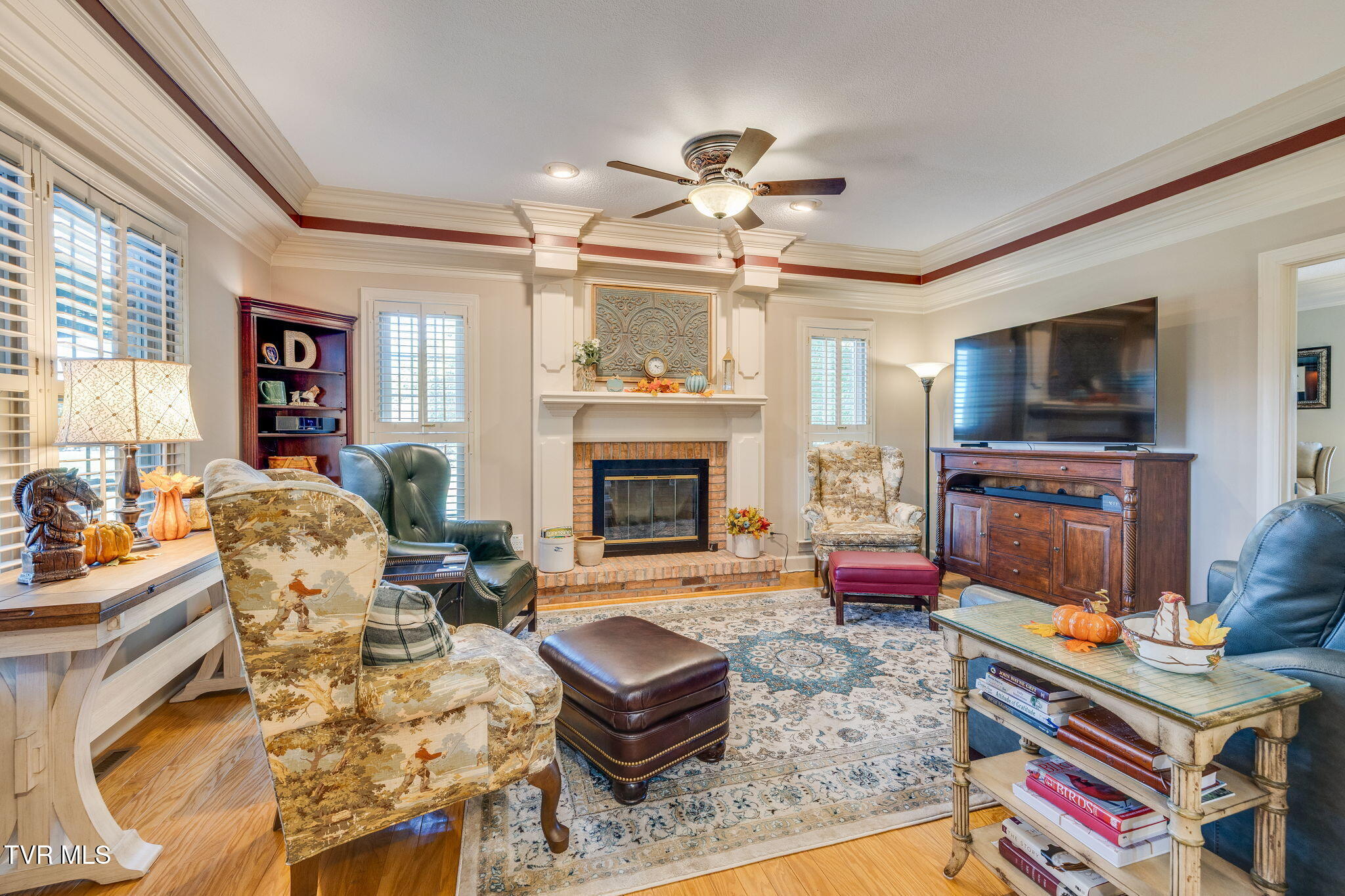 Furnished living room with a brick fireplace, ornate mantel, shuttered windows, and patterned rug.