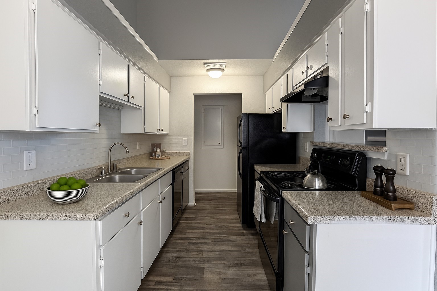 Galley kitchen with white cabinets, speckled counters, a double sink, black appliances, and dark wood-look flooring.
