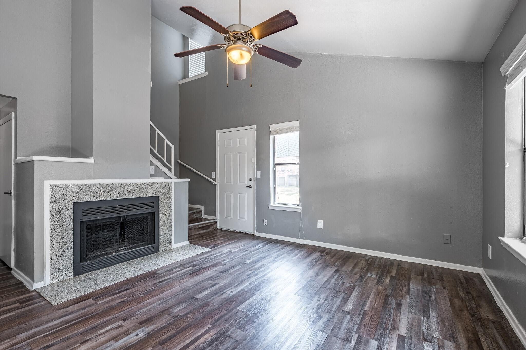 Empty living room with a speckled fireplace, dark flooring, and staircase by the front door.