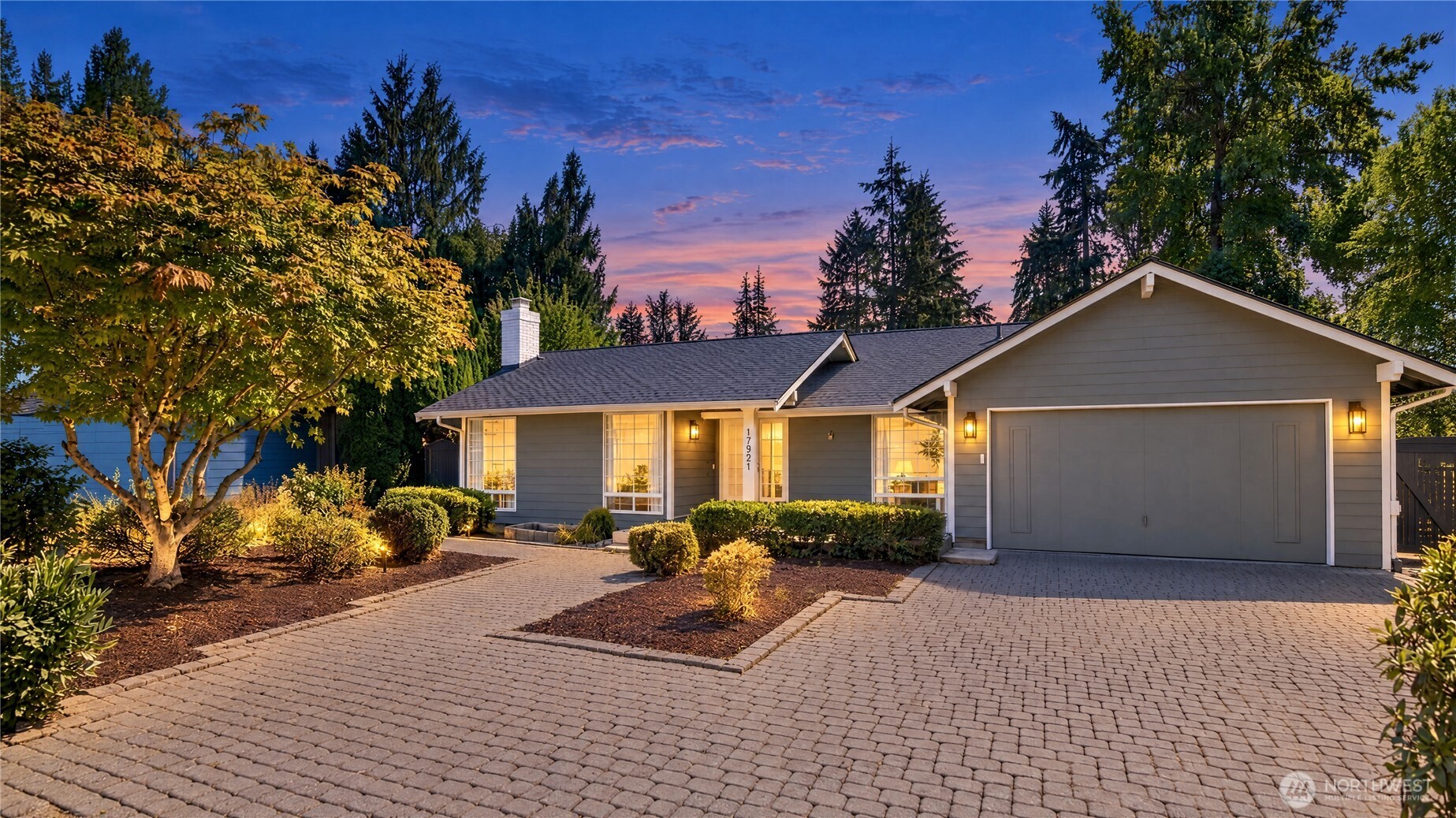 Single-story gray home with an attached garage and broad brick-paver driveway at dusk.