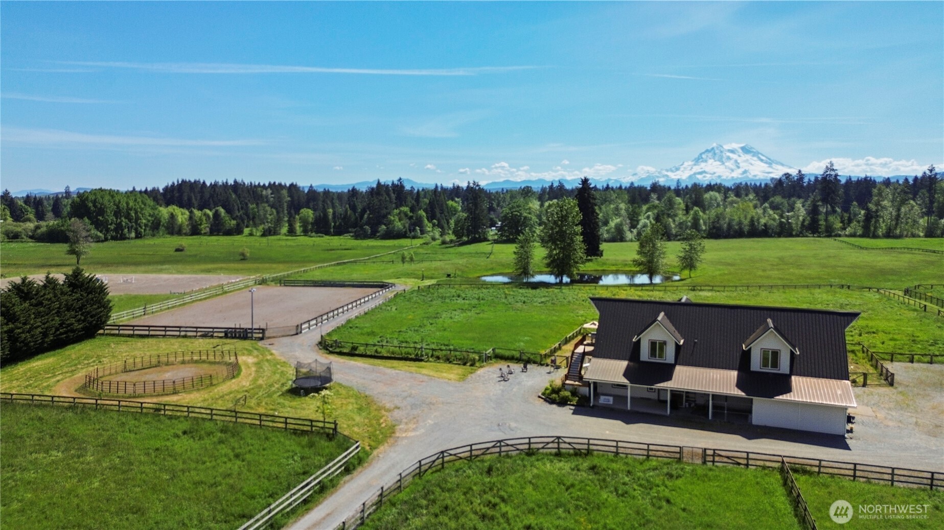 Aerial view of an equestrian property with fenced pastures, riding arenas, a pond, and a snow-capped mountain.
