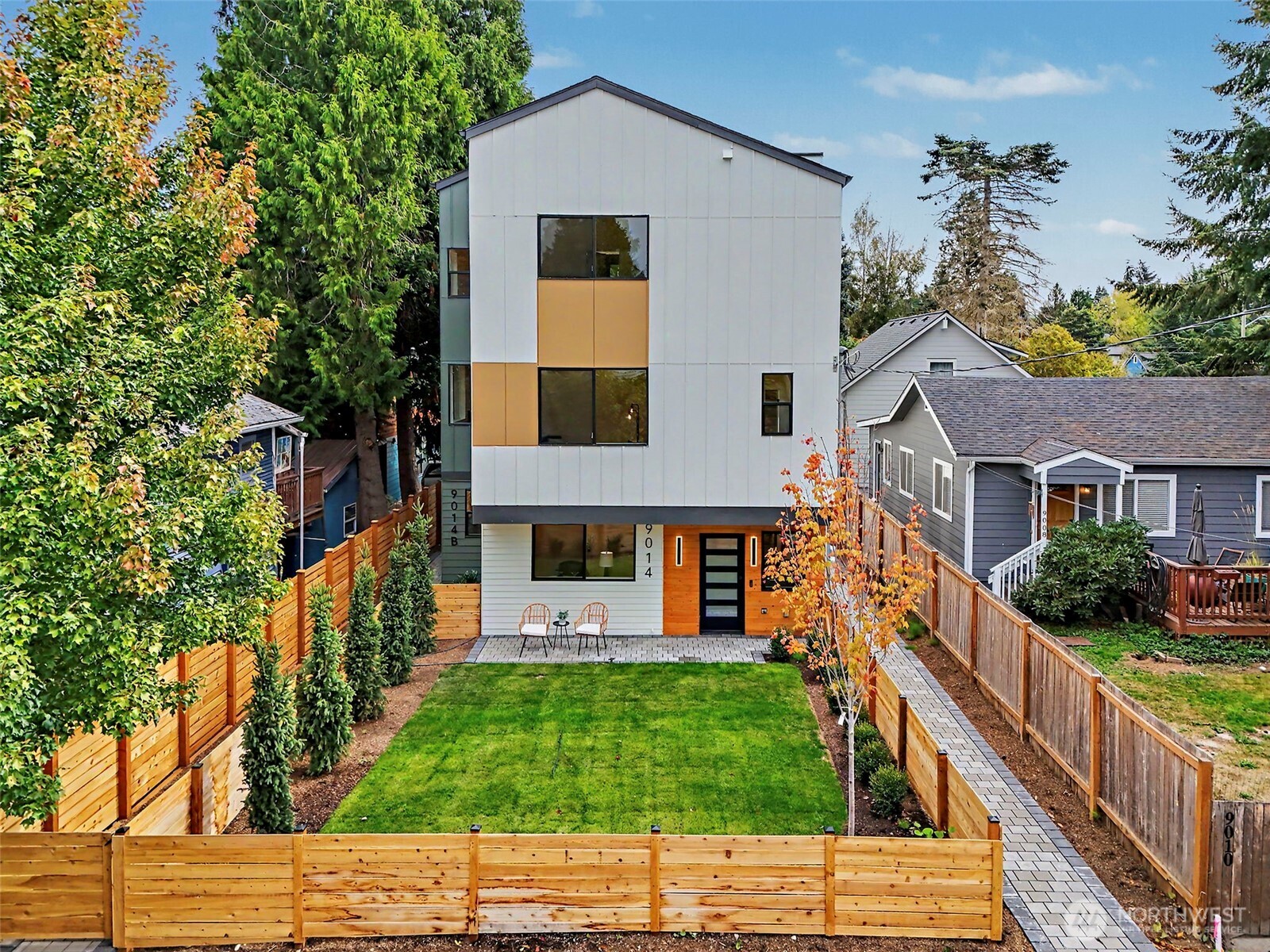 Modern three-story white house with wood accents, fenced lawn, paved walkway, and patio chairs.