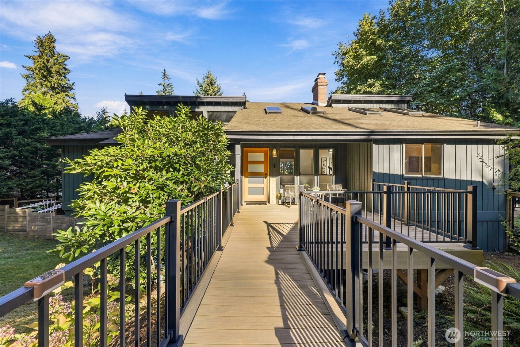 House entrance at the end of a long raised deck walkway, framed by mature trees.