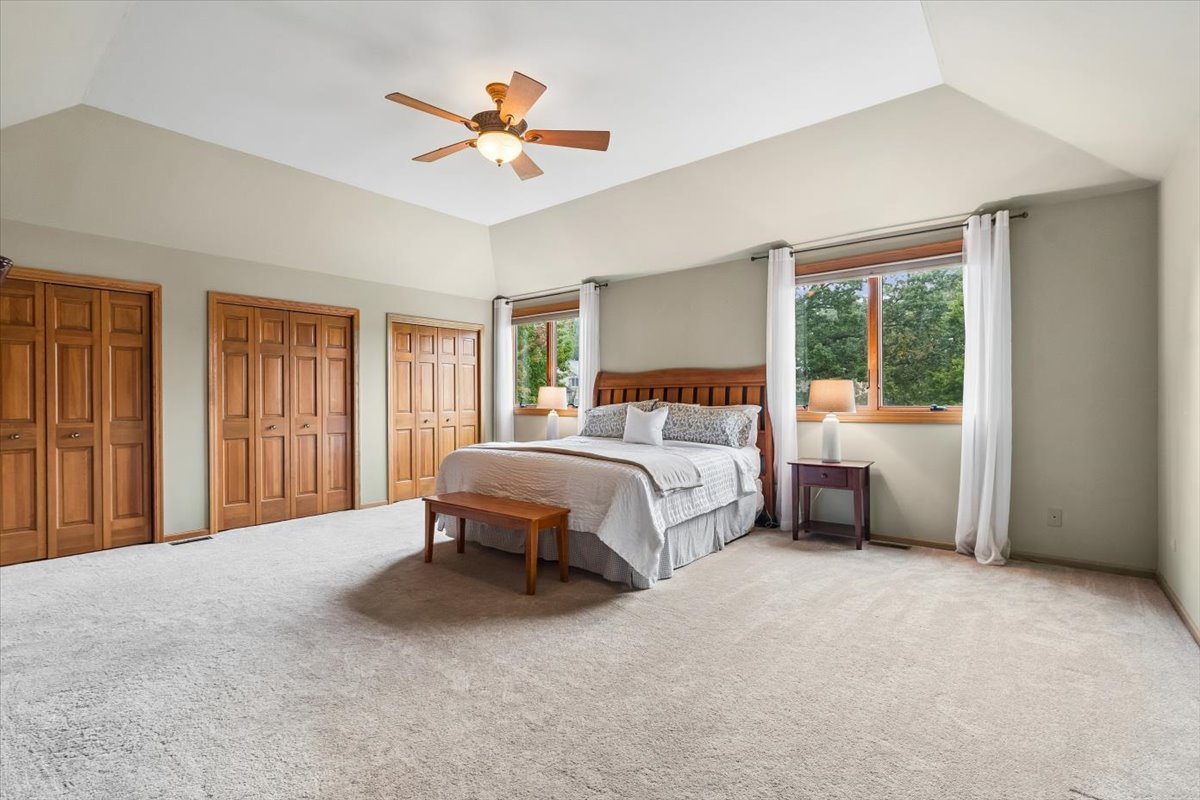 Spacious bedroom with wooden closets, a ceiling fan, and large windows overlooking trees.