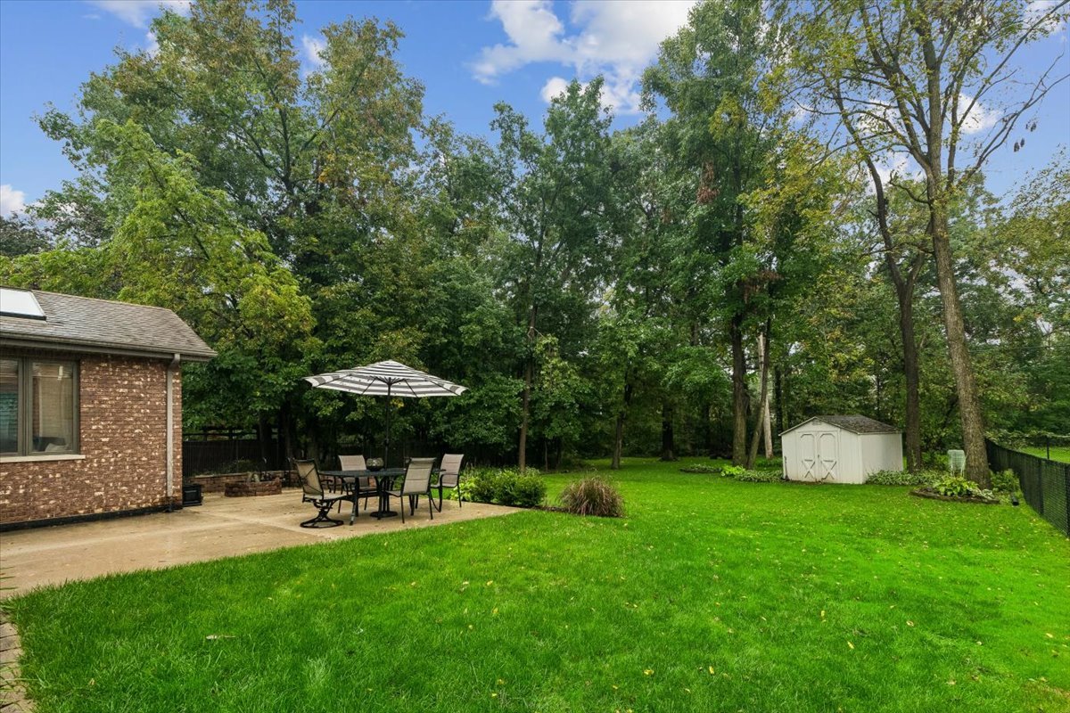 Backyard with a broad lawn, shaded patio dining set, mature trees, and a white storage shed.