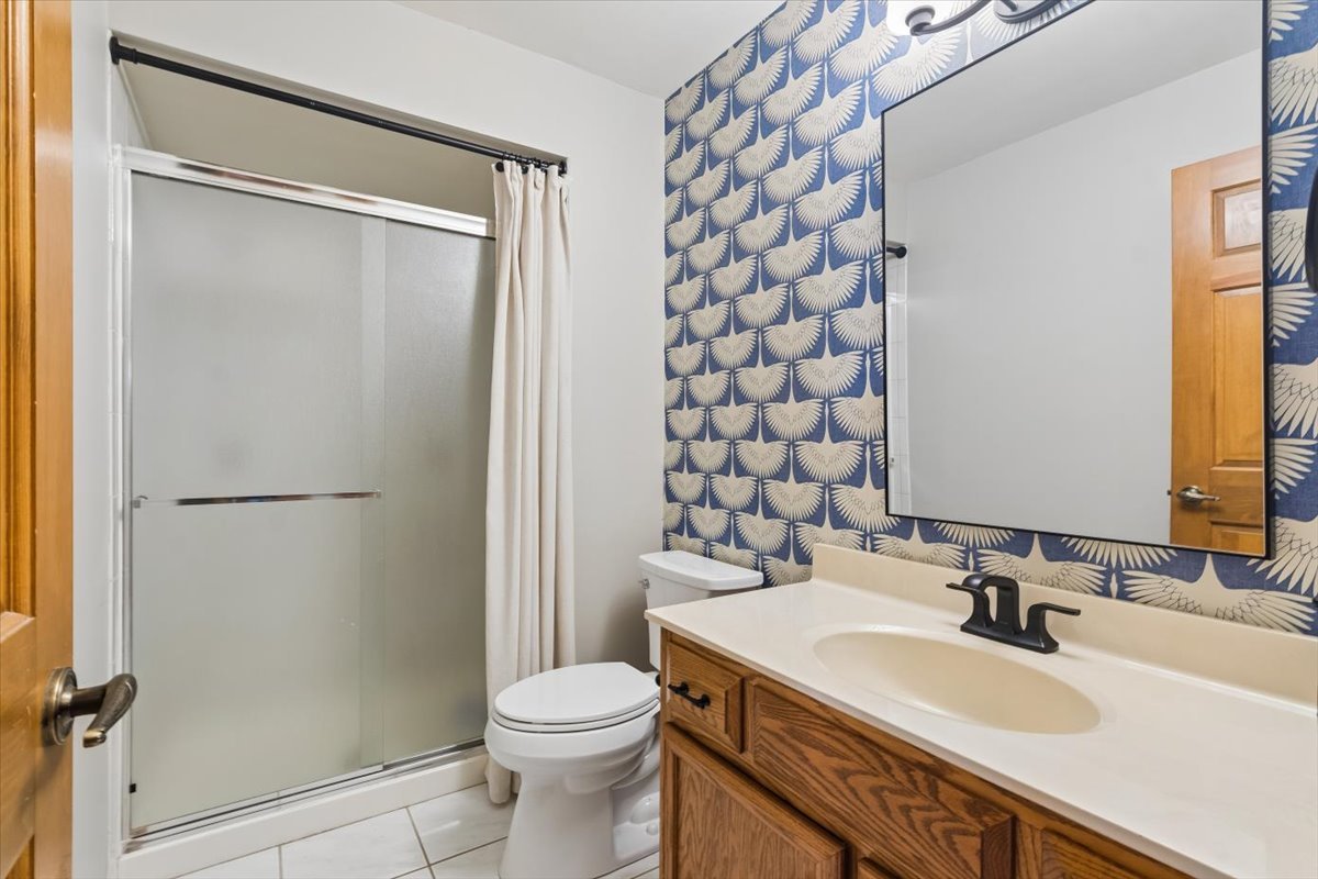 Bathroom with glass shower, wood vanity, and blue-and-cream patterned wallpaper.