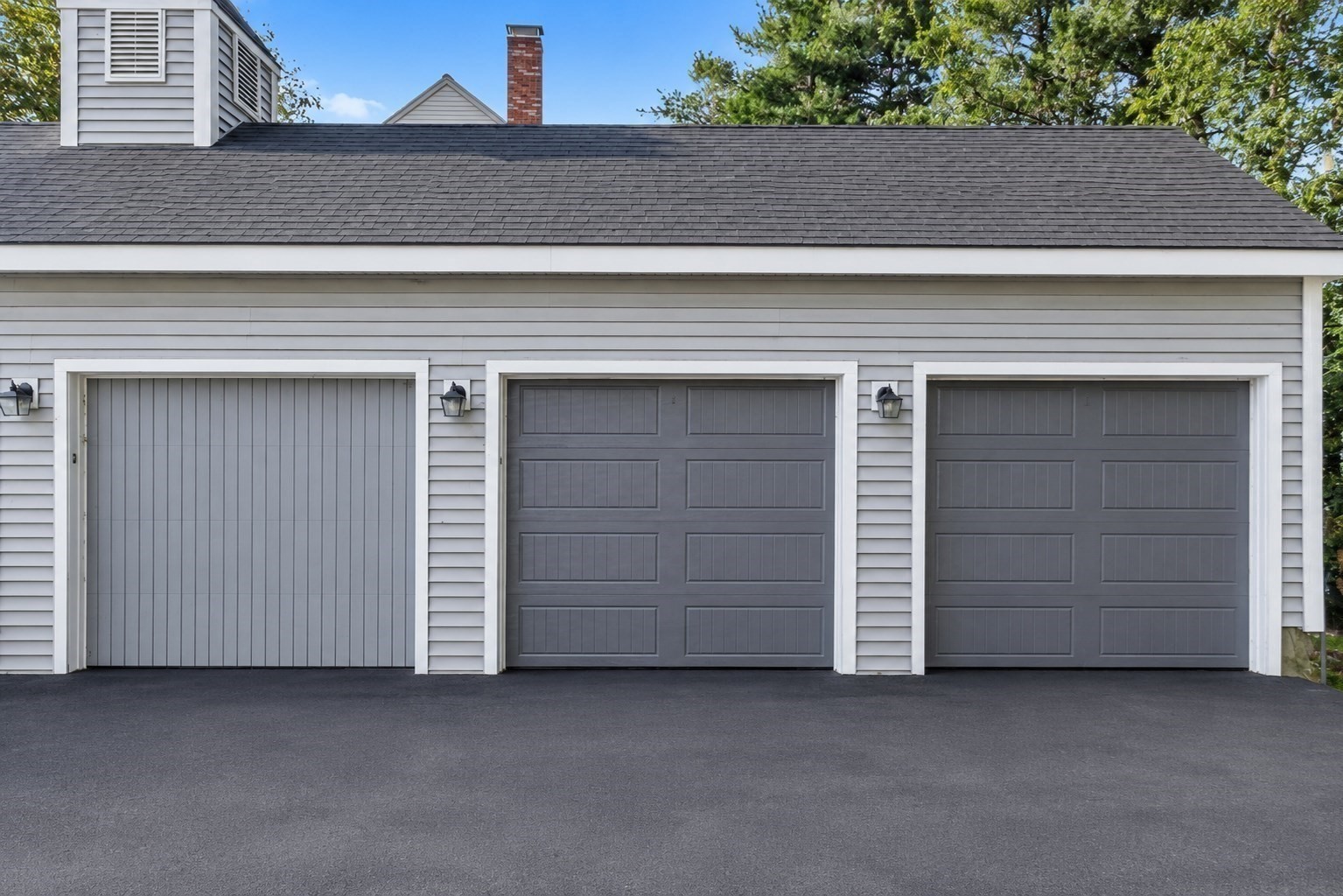 Gray detached garage with three doors, white trim, and a wide asphalt driveway.