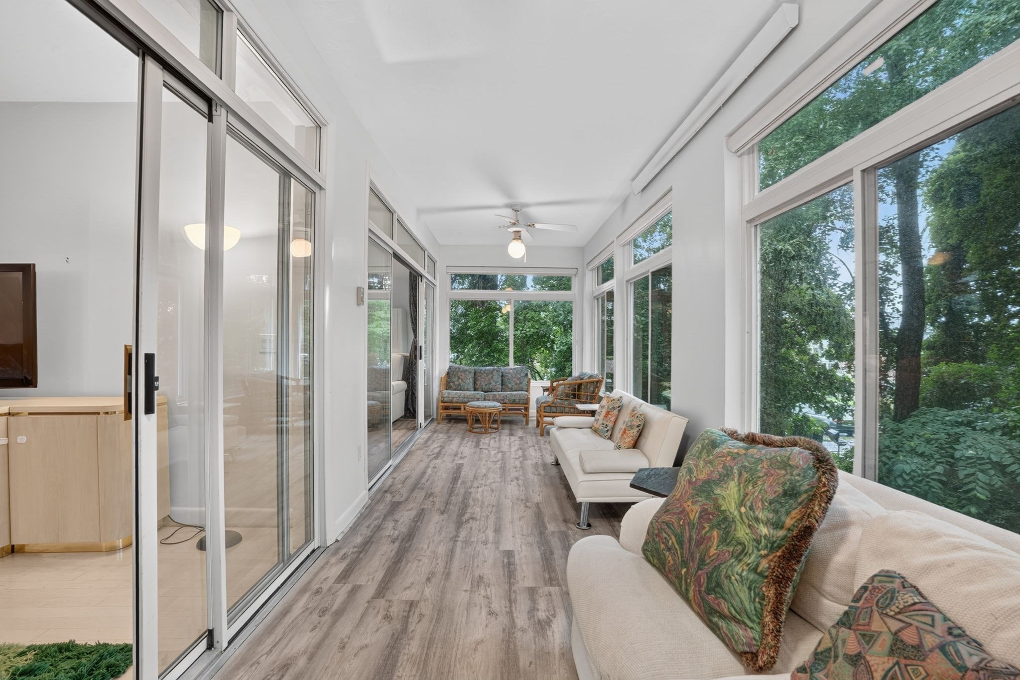 Sunroom with white sofas, wicker seating, and large windows overlooking leafy trees.