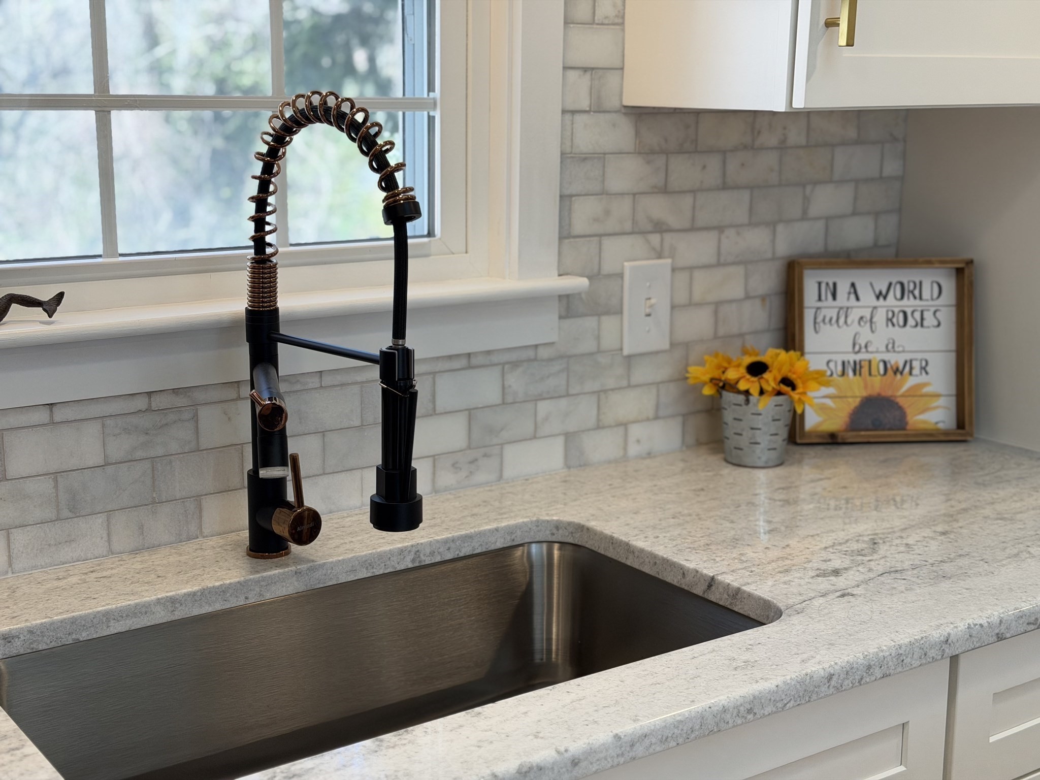 Kitchen sink with a black spring-coil faucet, pale stone counter, gray tile backsplash, and sunflowers.