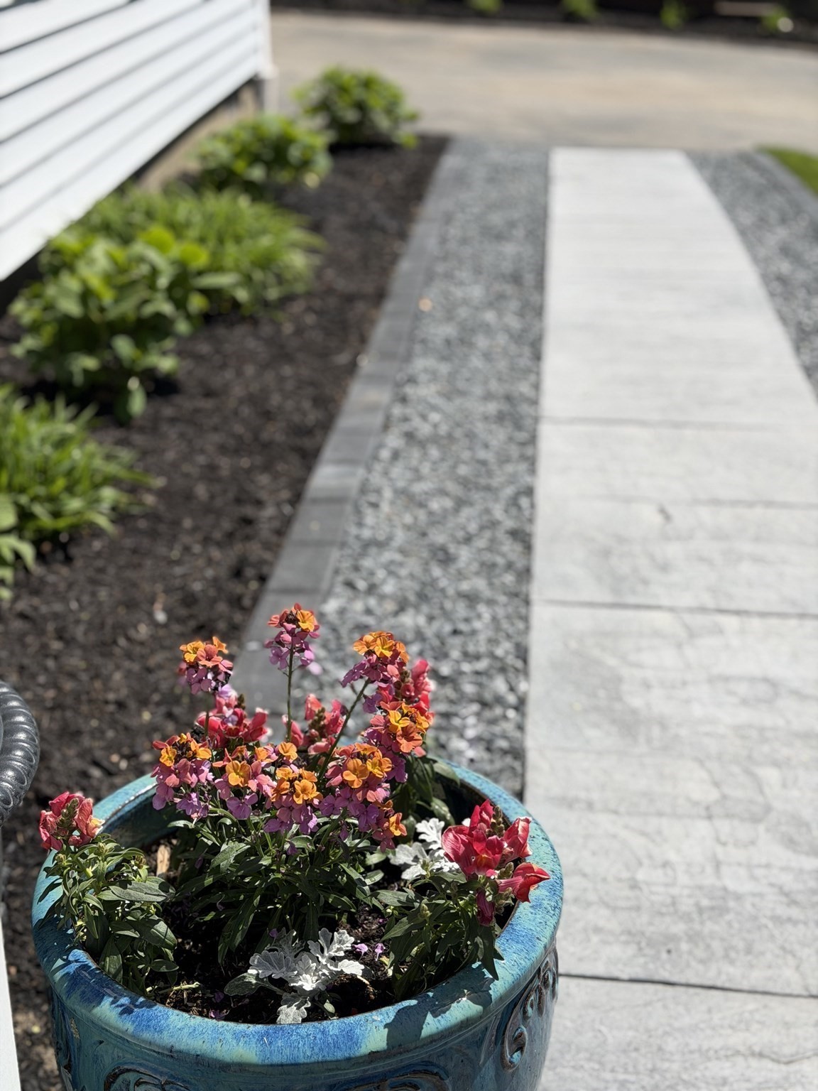 Colorful flowers in a blue planter beside a paved walkway and gravel path.