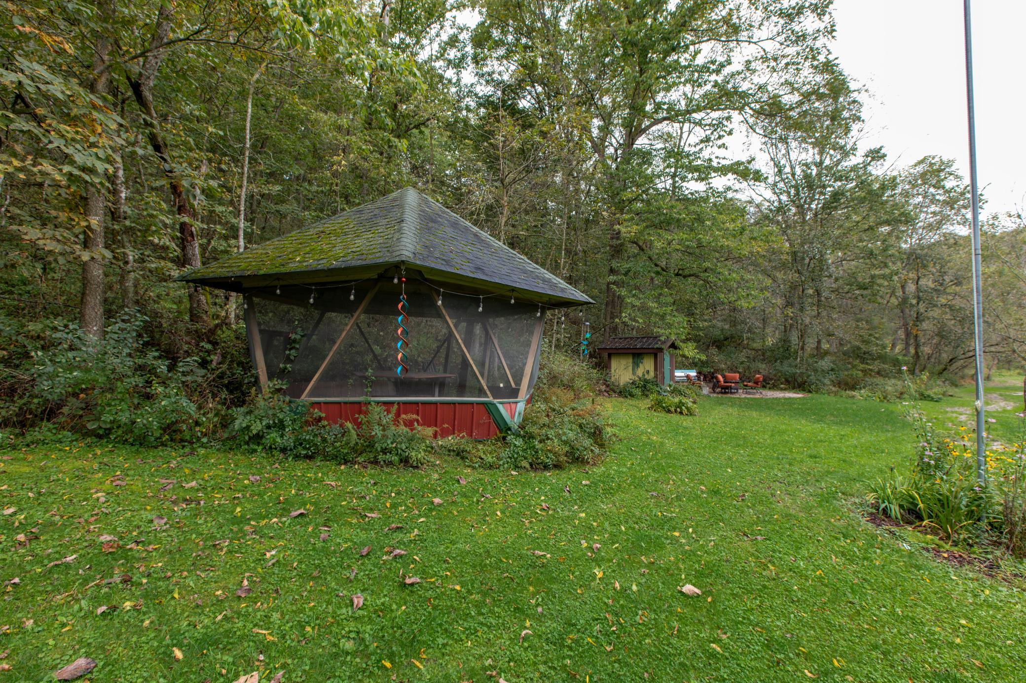 Screened gazebo on a grassy lawn beside woods, with a small shed and outdoor seating nearby.