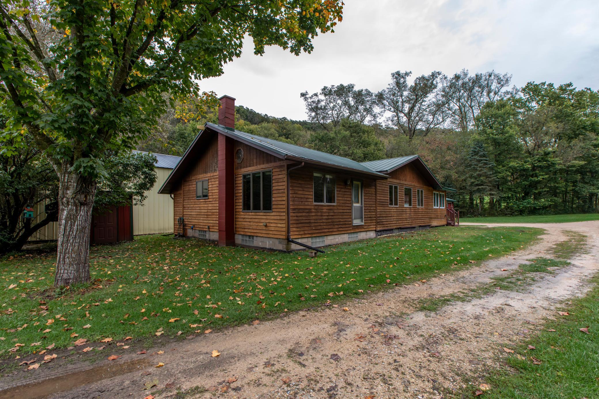 Wood-sided home with a metal roof and red chimney beside a gravel driveway and wooded yard.