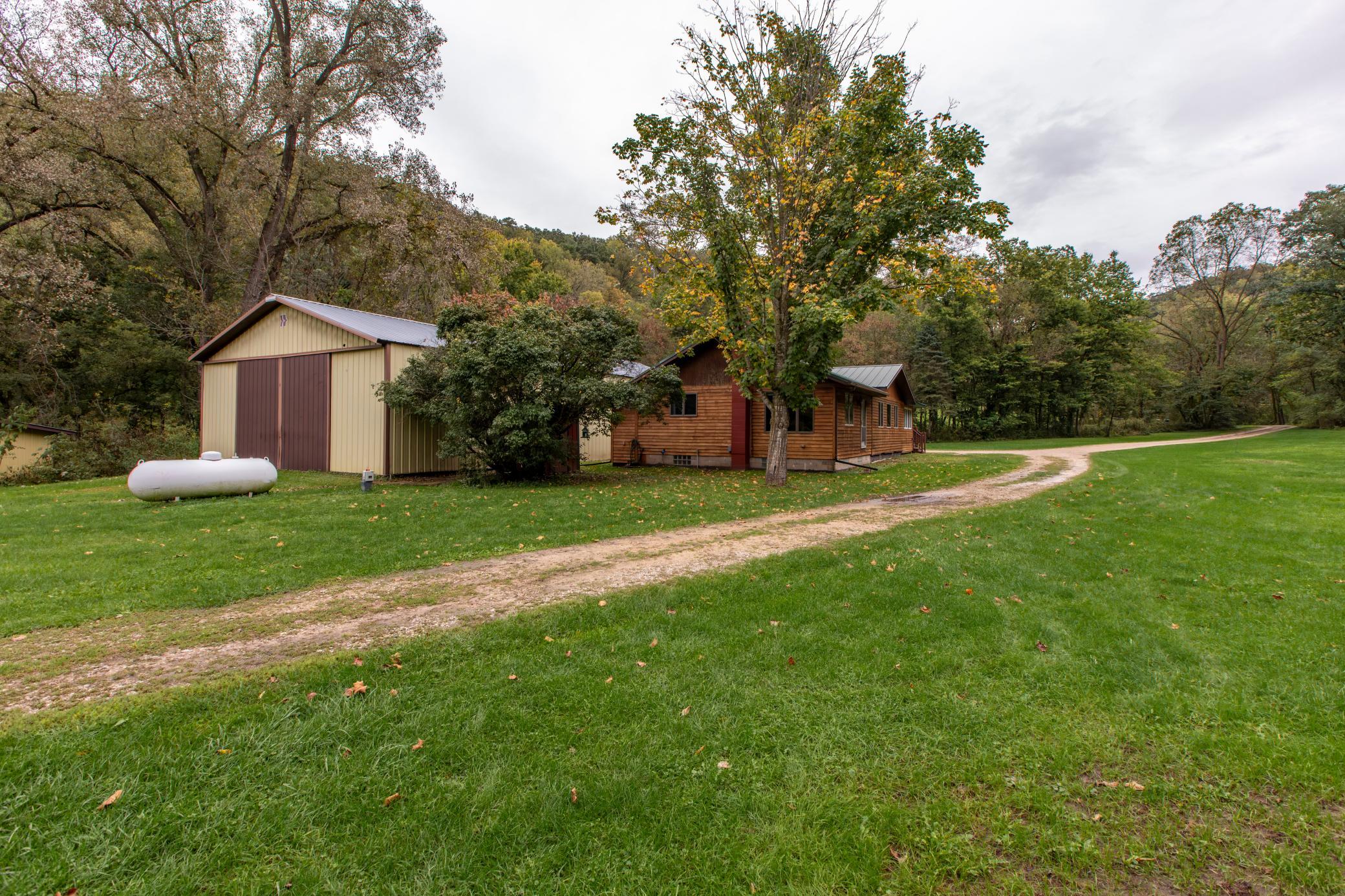 Wood-sided house and large outbuilding beside a curving gravel drive on a wooded rural property.