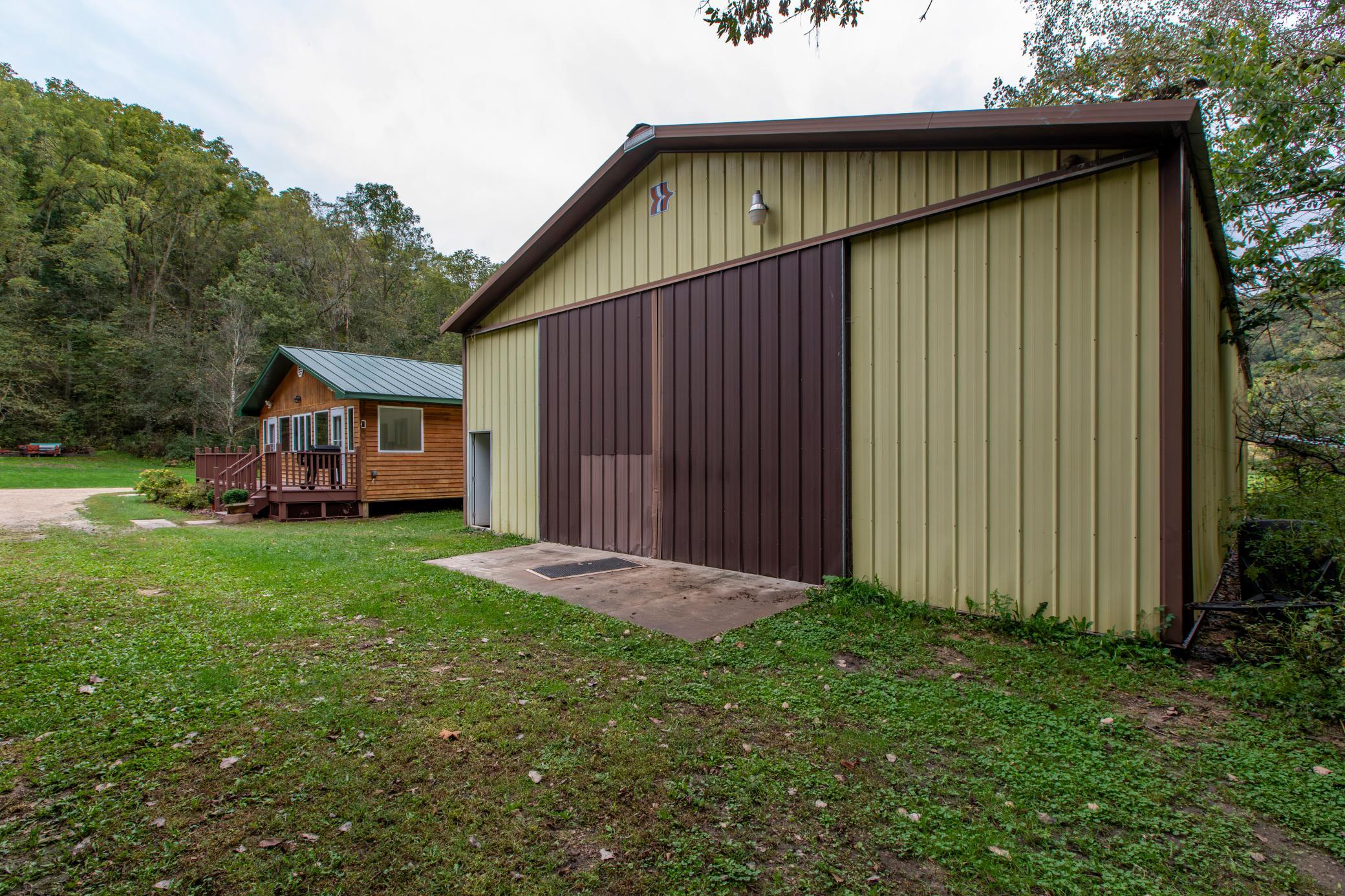 Large metal-sided barn beside a small wood cabin on a grassy, wooded property.