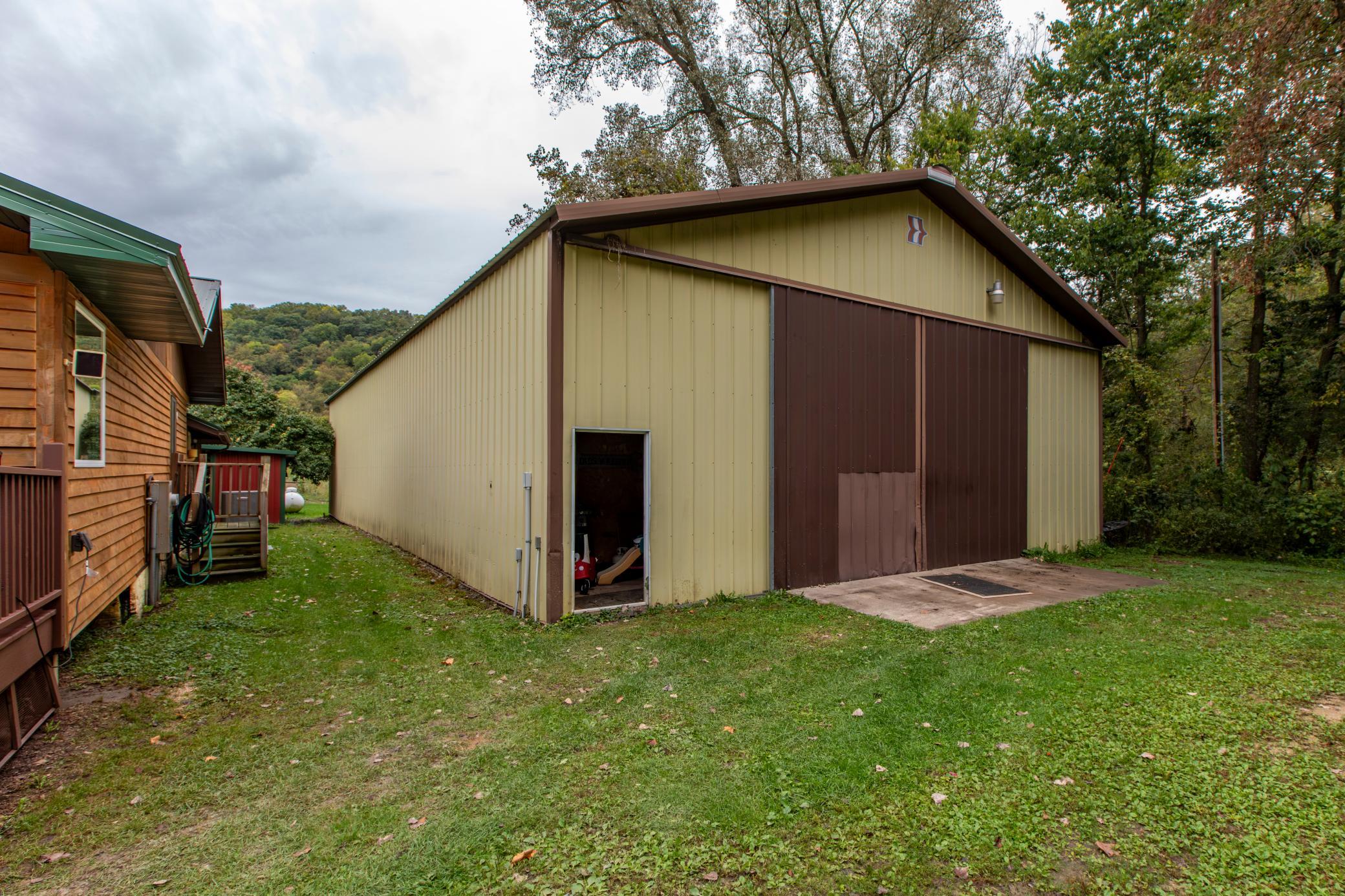 Large pale yellow metal outbuilding with brown sliding doors beside a wood-sided house and wooded yard.