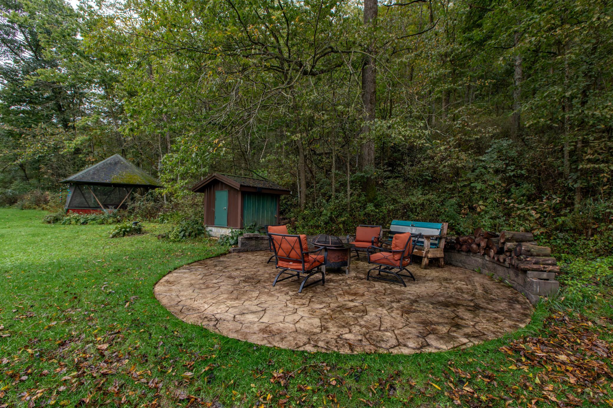 Backyard patio with orange chairs around a fire pit, a small shed, and a gazebo among the trees.