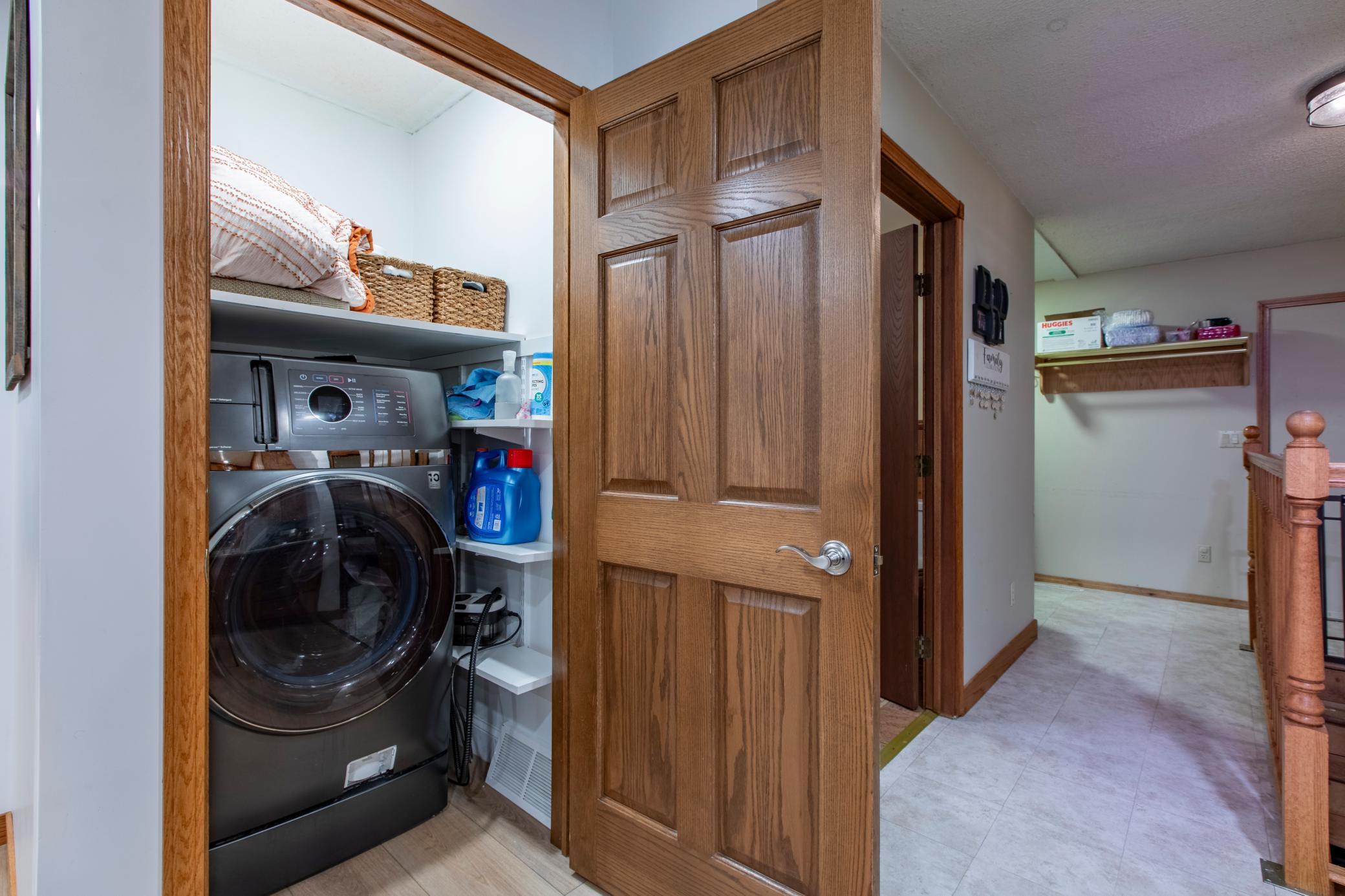 Laundry closet with a front-loading washer and dryer, open wooden door, and shelves of supplies.