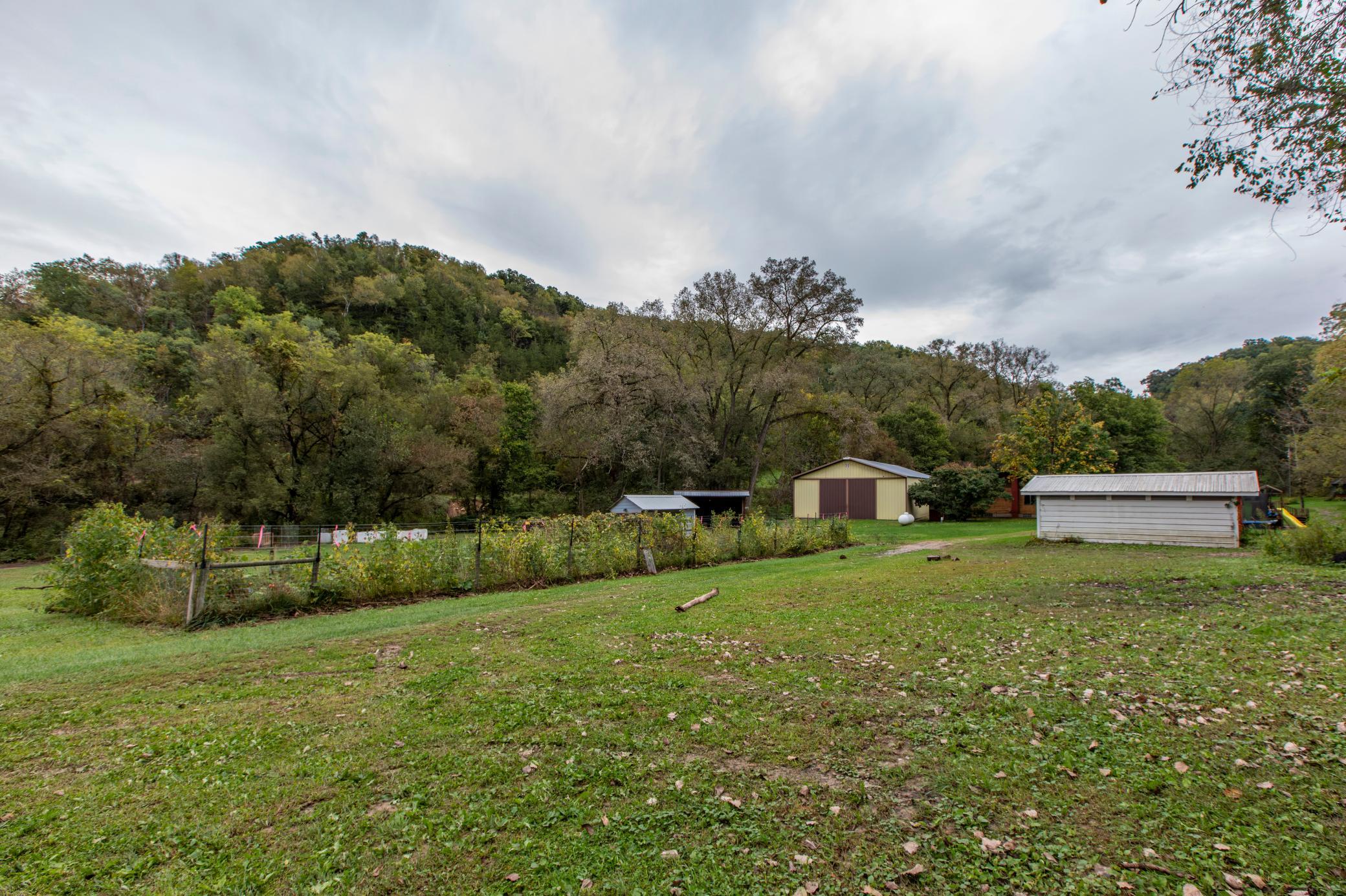 Rural yard with a fenced garden and several outbuildings below wooded hills.