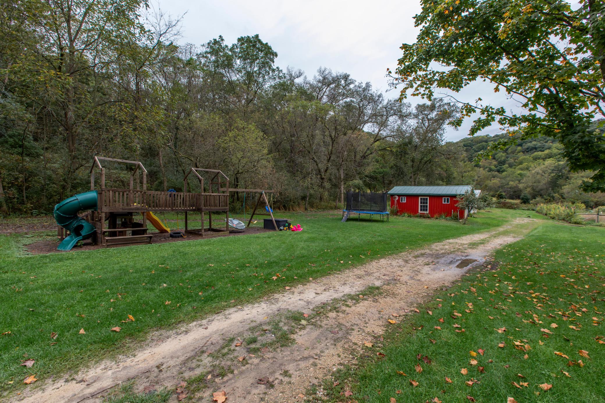 Rural yard with a wooden playset, trampoline, and red outbuilding beside wooded hills.
