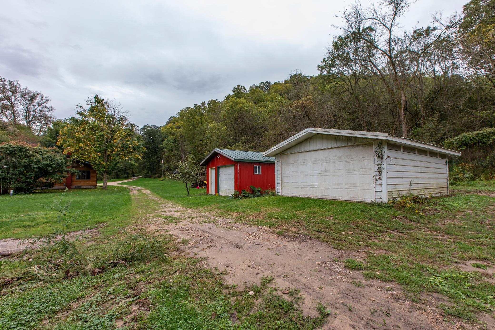 White detached garage and red outbuilding beside a gravel driveway, with woods behind.