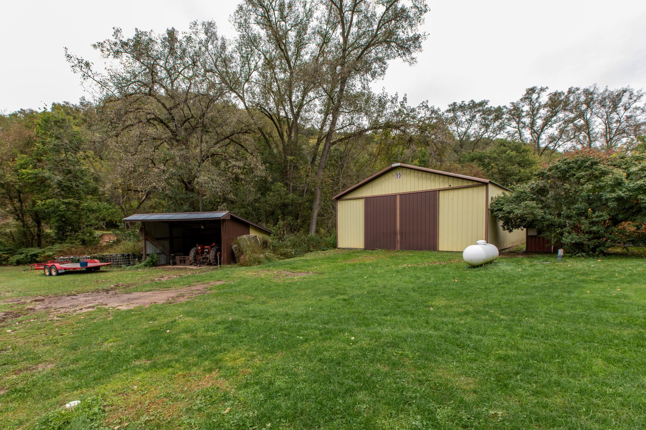 Yellow metal storage barn and open equipment shed beside a grassy yard and wooded area.
